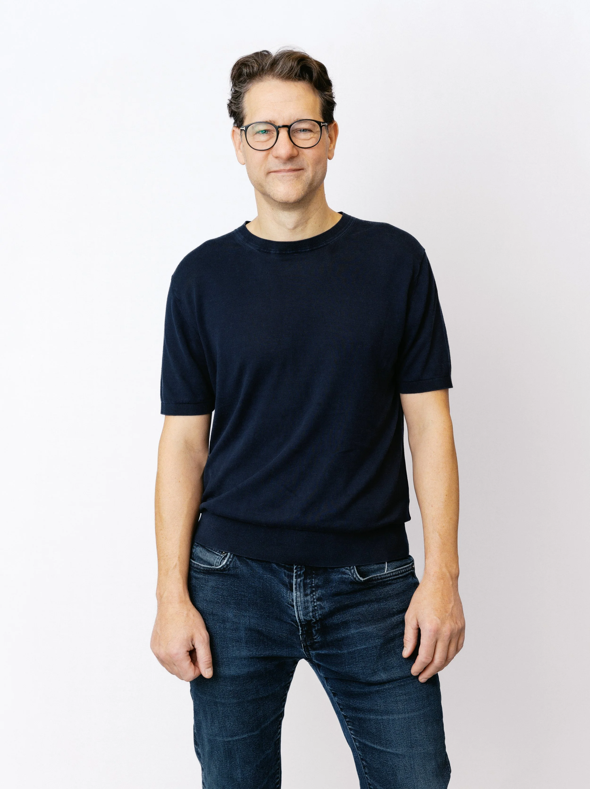 En man med glasögon och mörk vågig hår, bär en svart T-shirt och jeans, står mot en vit bakgrund.