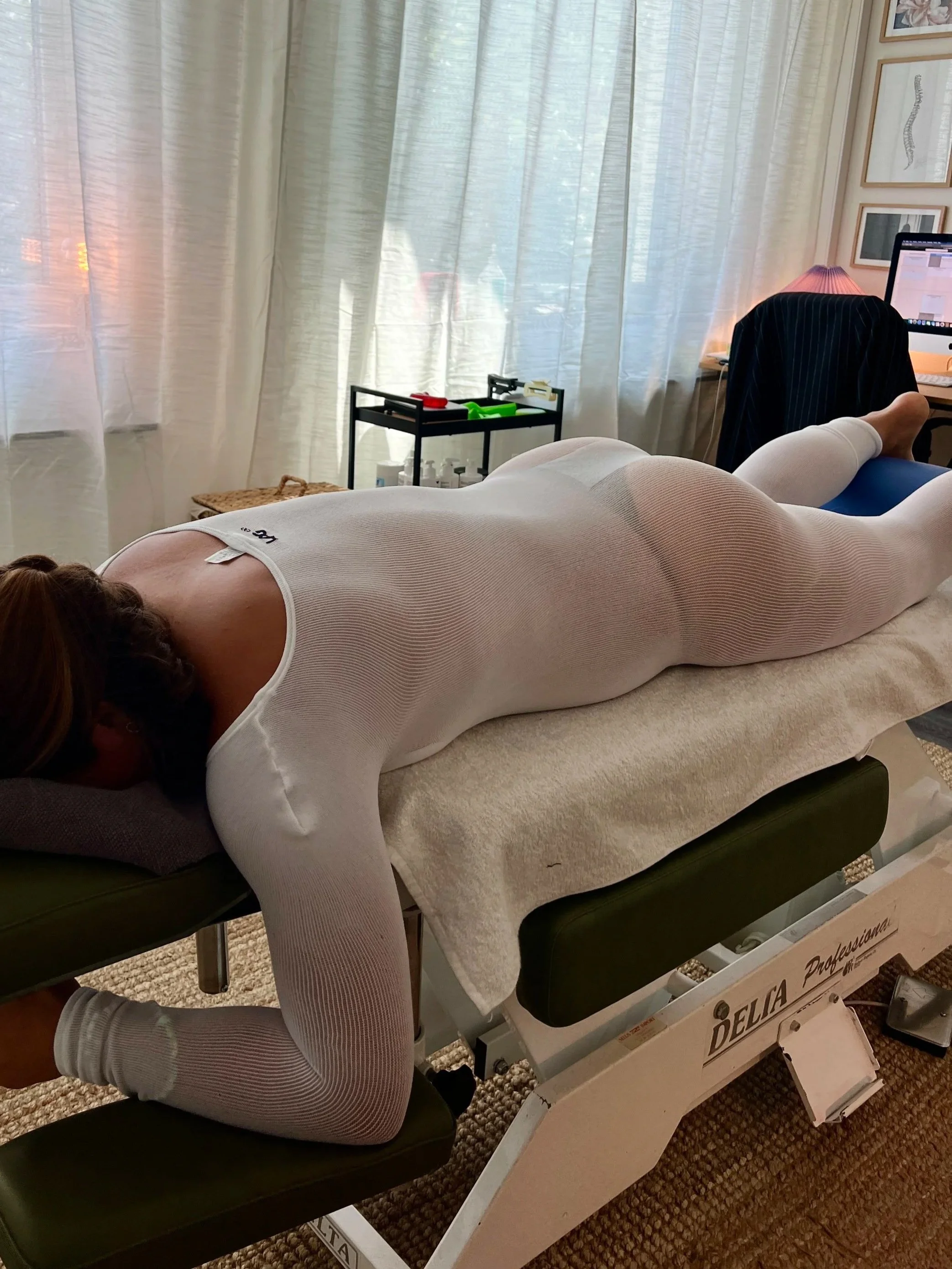 En person ligger på en massagebänk i ett rum, med en person i vit kroppsstrumpa