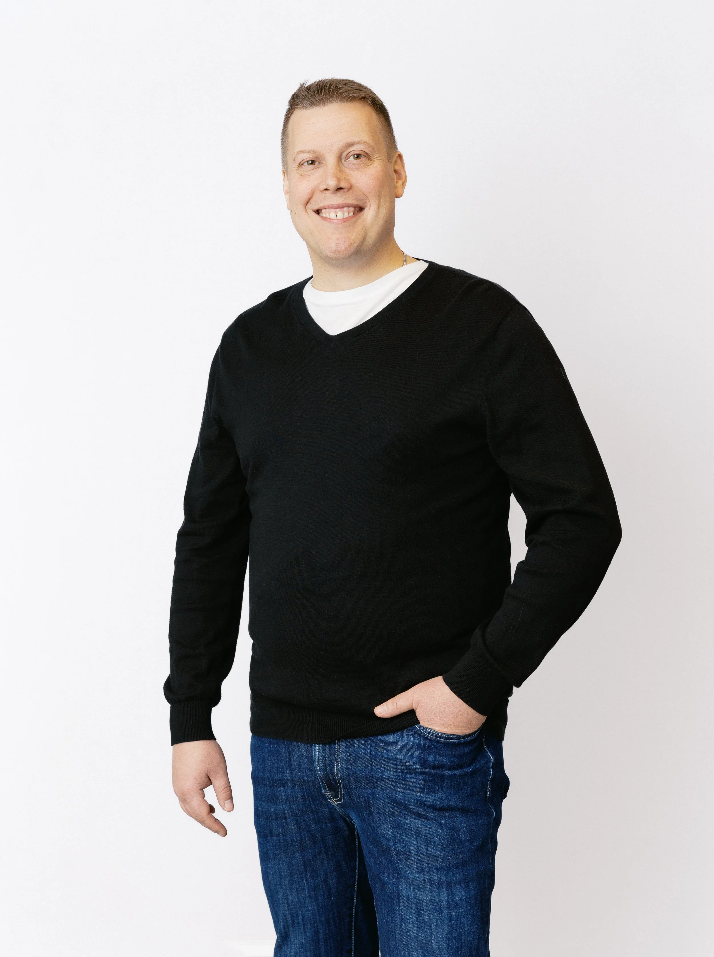 En man med kort hår, helskägg, svart tröja och blå jeans som står mot en vit bakgrund.