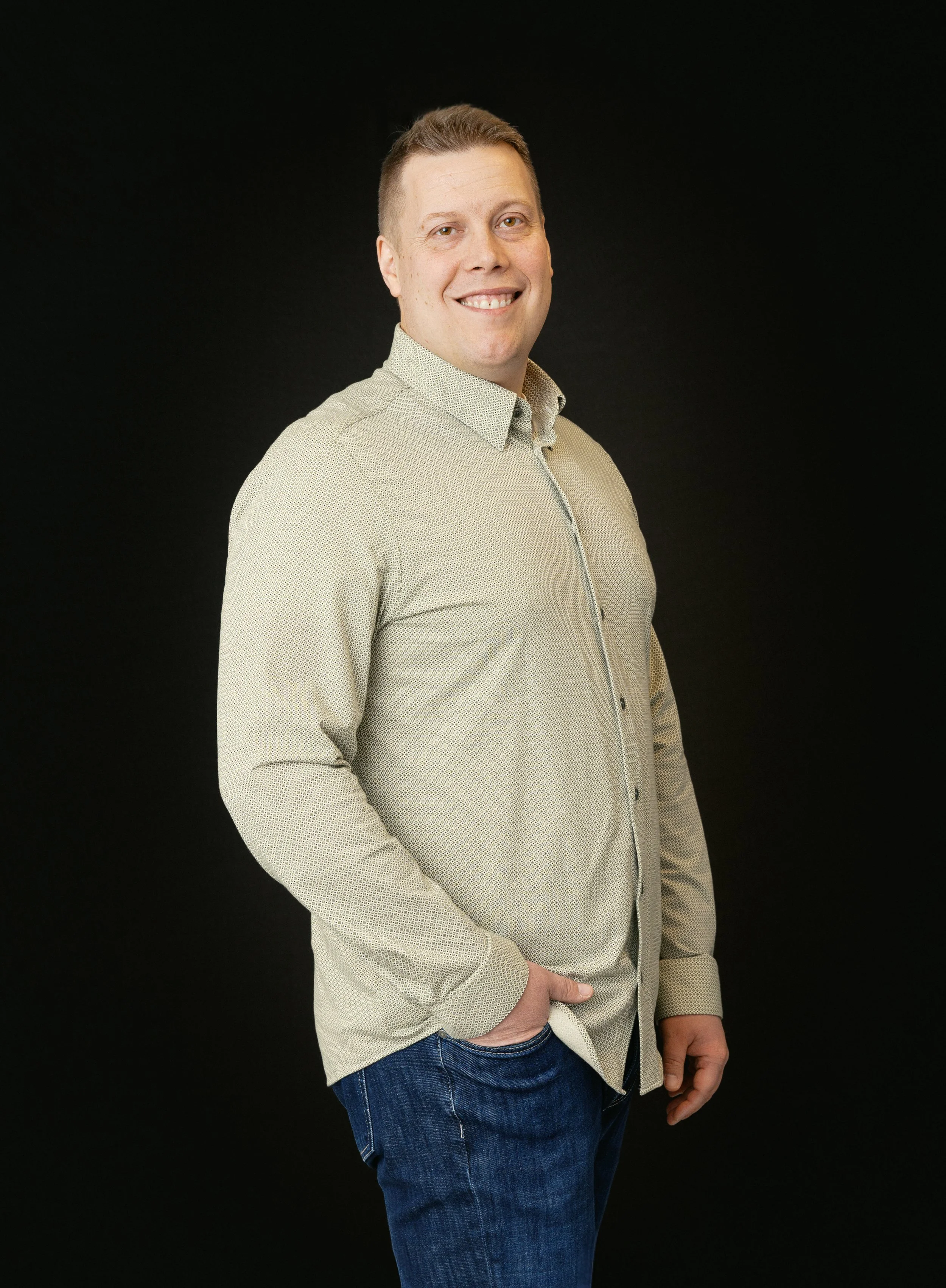 Man i beige skjorta och blå jeans står mot svart bakgrund, ler och har handen i byxfickan.