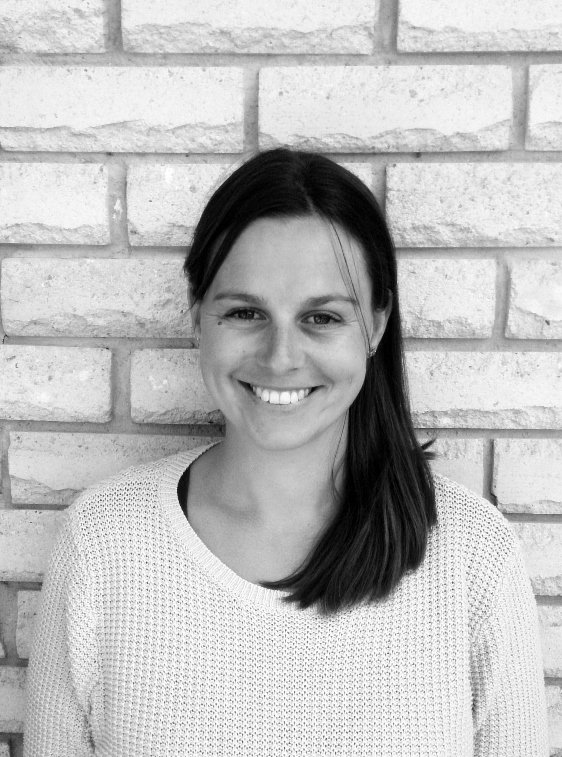 A woman with long dark hair smiling, wearing a light-colored knitted sweater, standing against a light brick wall.