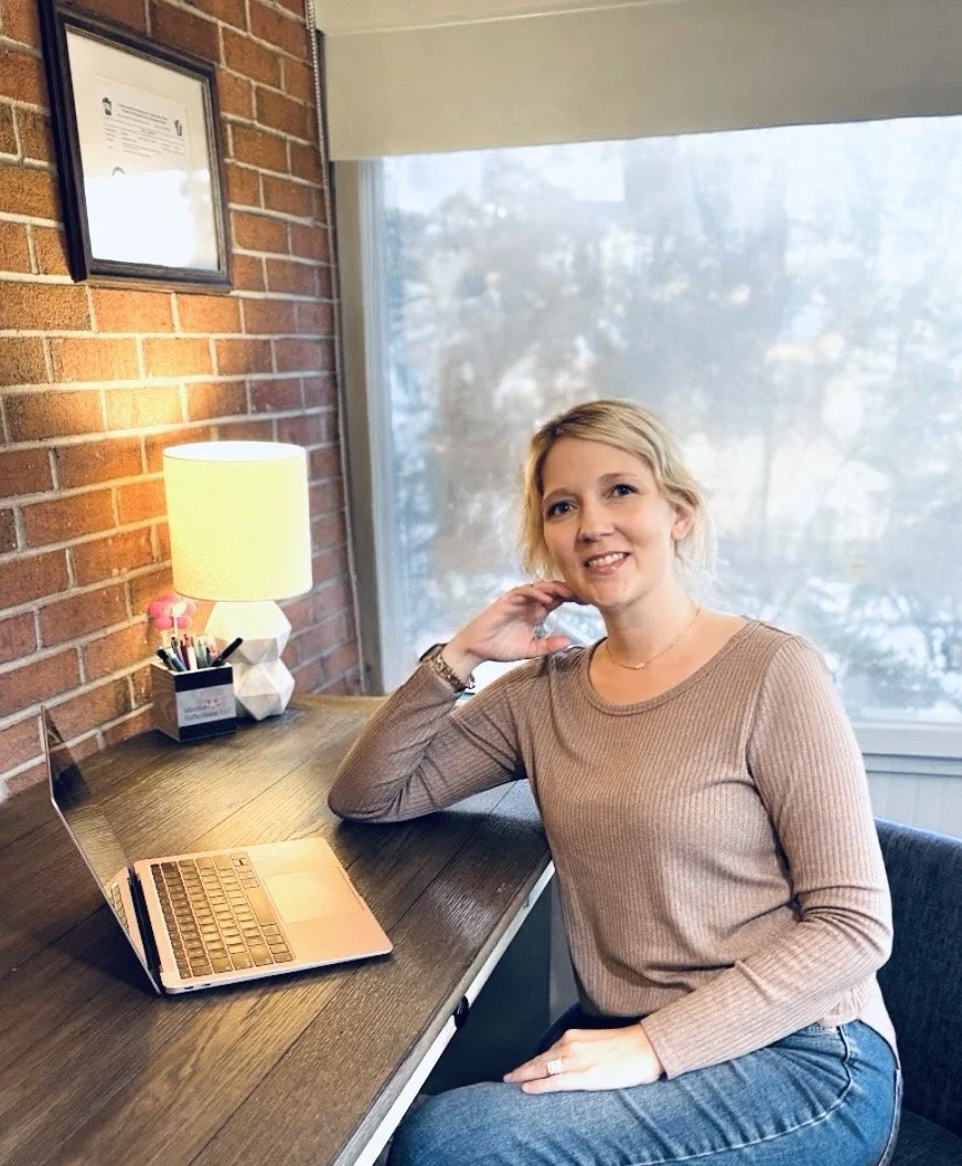 A woman with blonde hair sits at a wooden desk with a laptop, a desk lamp, a container of pens and pencils, and a certificate or diploma on the brick wall. She is smiling and wearing a tan long-sleeve top and blue jeans, next to a window with a view of trees outside.
