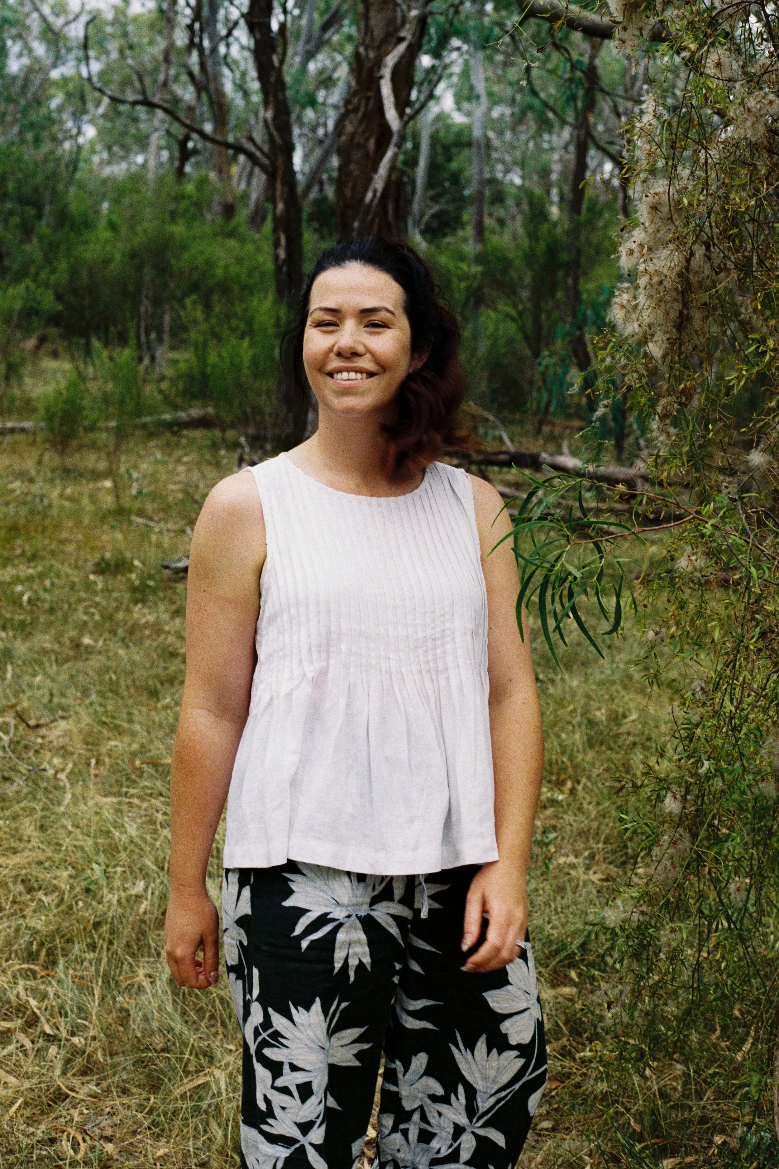 A woman standing outdoors in a forest or wooded area with trees and greenery, smiling at the camera, wearing a white sleeveless top and black pants with a white floral pattern.