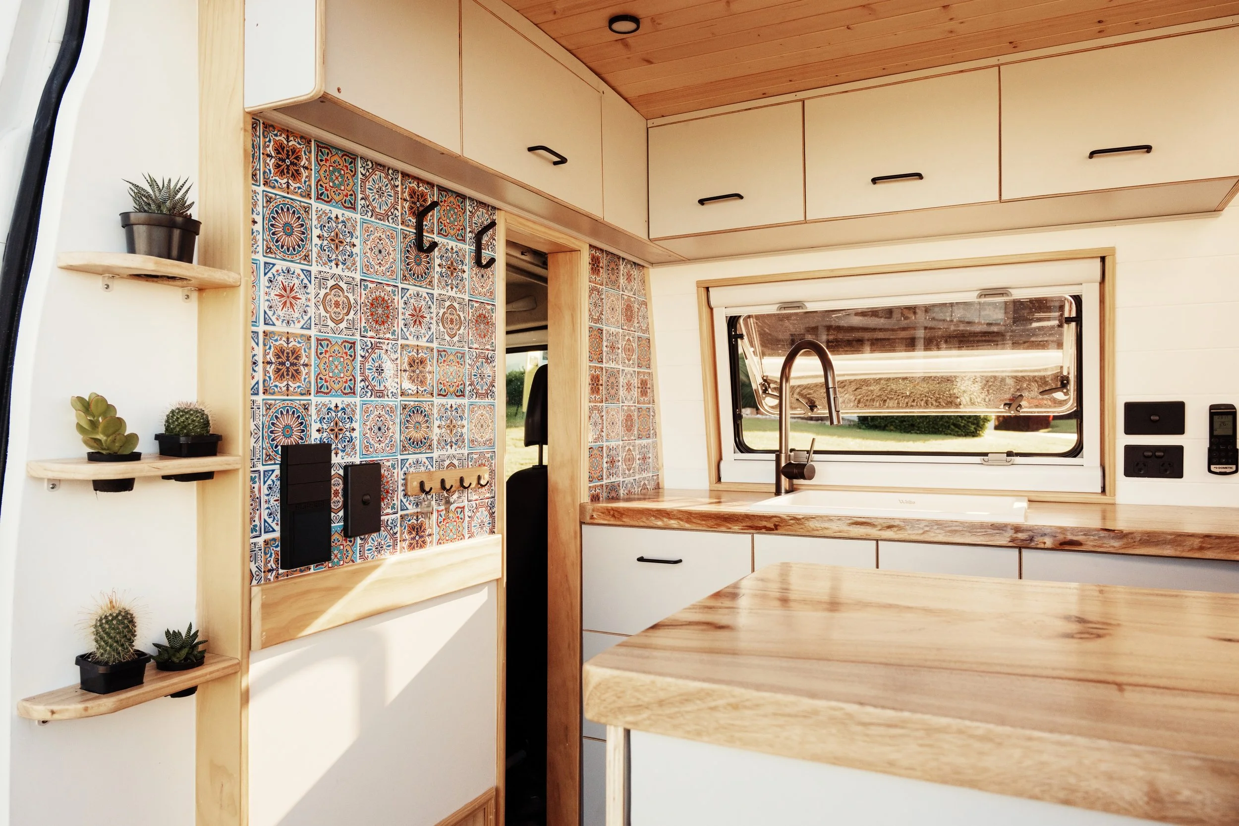 Interior of a tiny home kitchen with wooden cabinets, a window, patterned tile wall, and small shelves with potted plants.