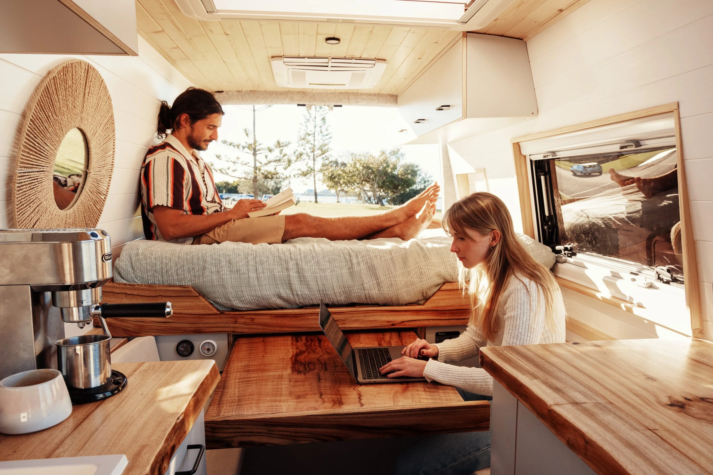 Interior of a camper van with a young man lying on a bed reading a book and a young woman working on a laptop at a wooden table, sunlight streaming in through an open window.