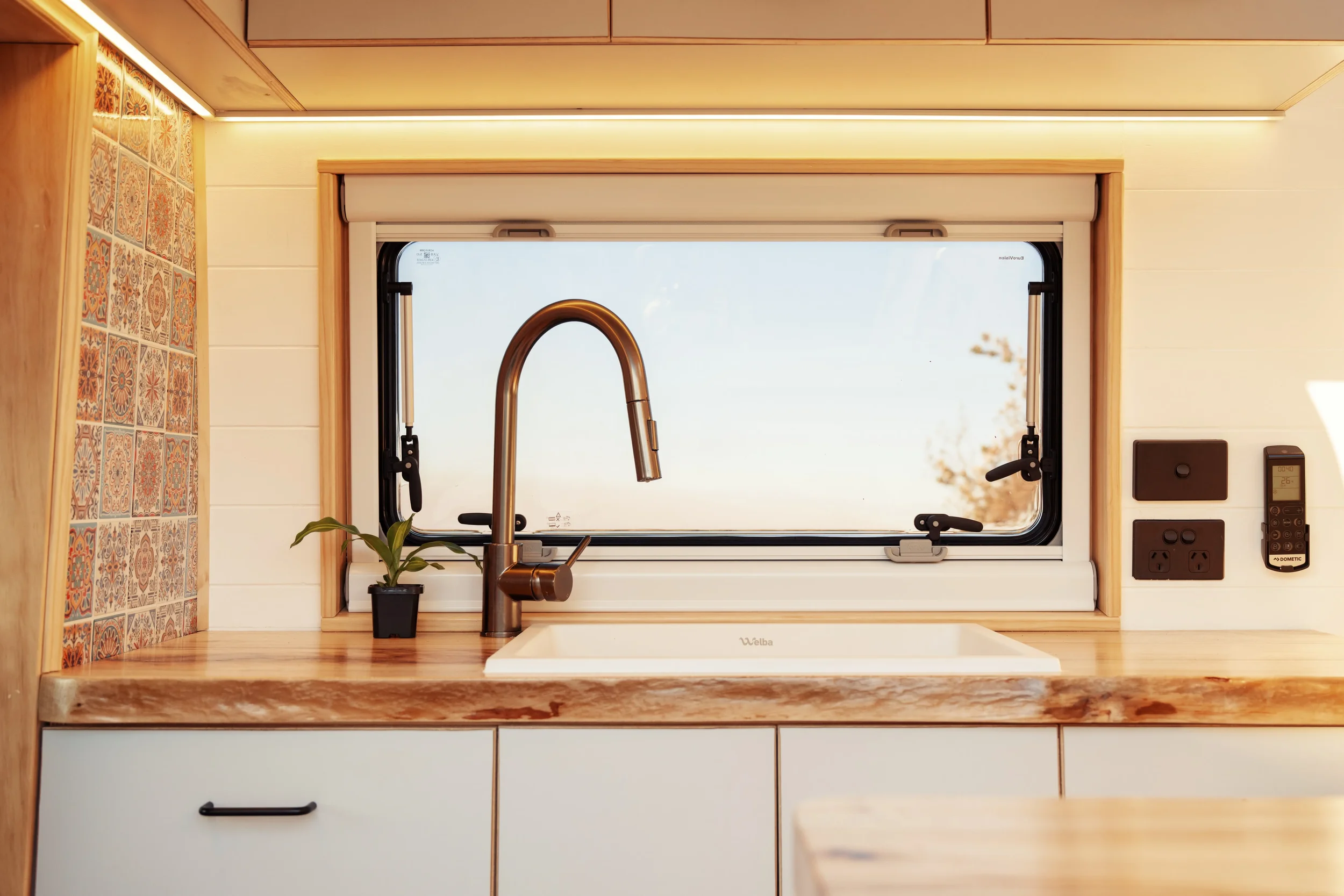 A kitchen countertop with a sink and a brass-colored faucet, a small potted plant, a window behind the sink, and various electrical outlets on the wall.