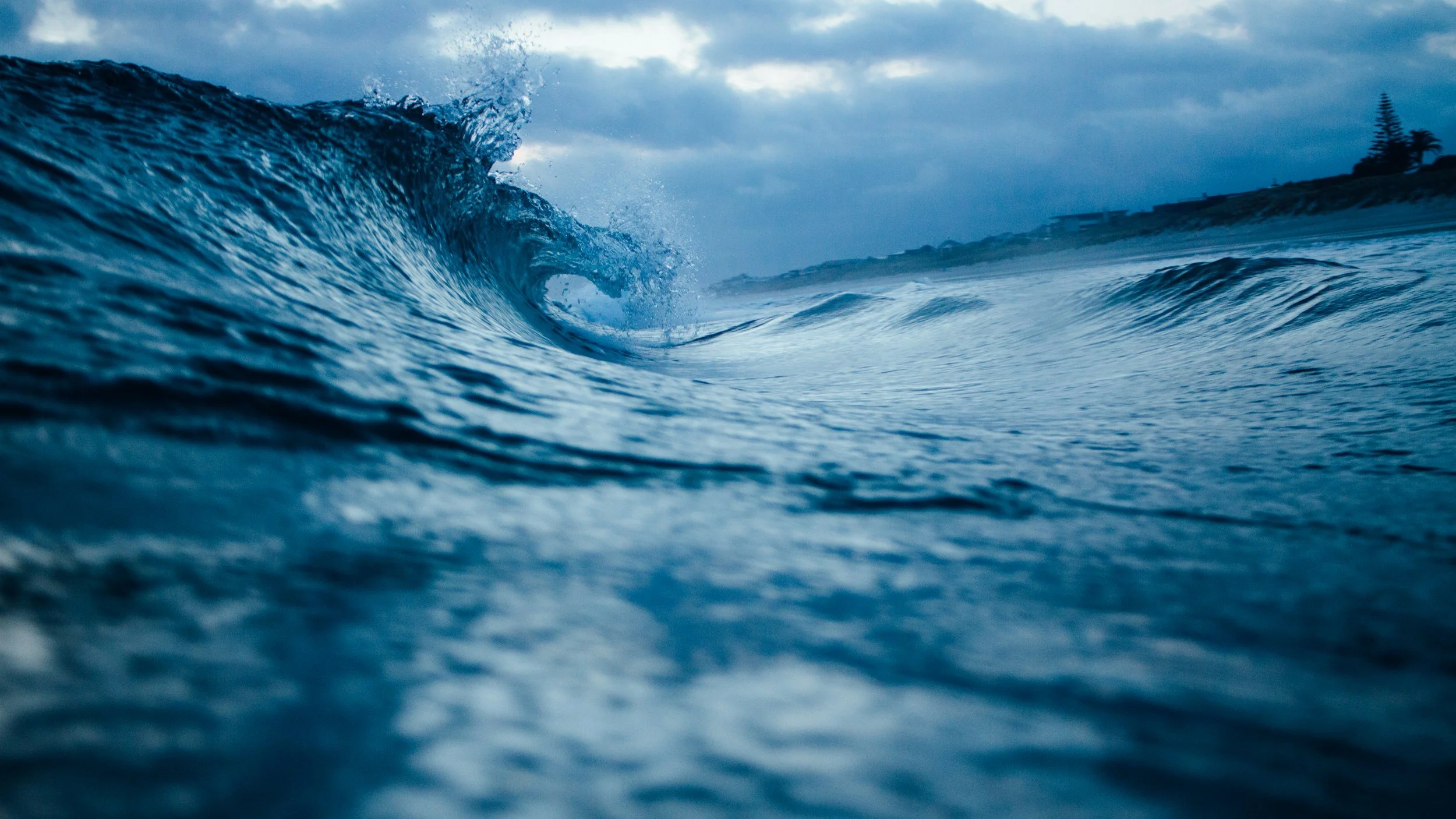 Eine große Welle im Meer mit einer Küstenlinie im Hintergrund bei bewölktem Himmel.