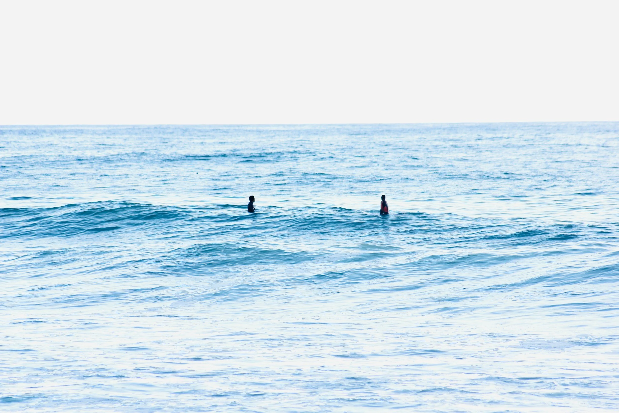 Zwei Kinder stehen im Wasser am Strand, mit leichtem Wellengang um sie herum, während der Horizont und der Himmel im Hintergrund sichtbar sind.
