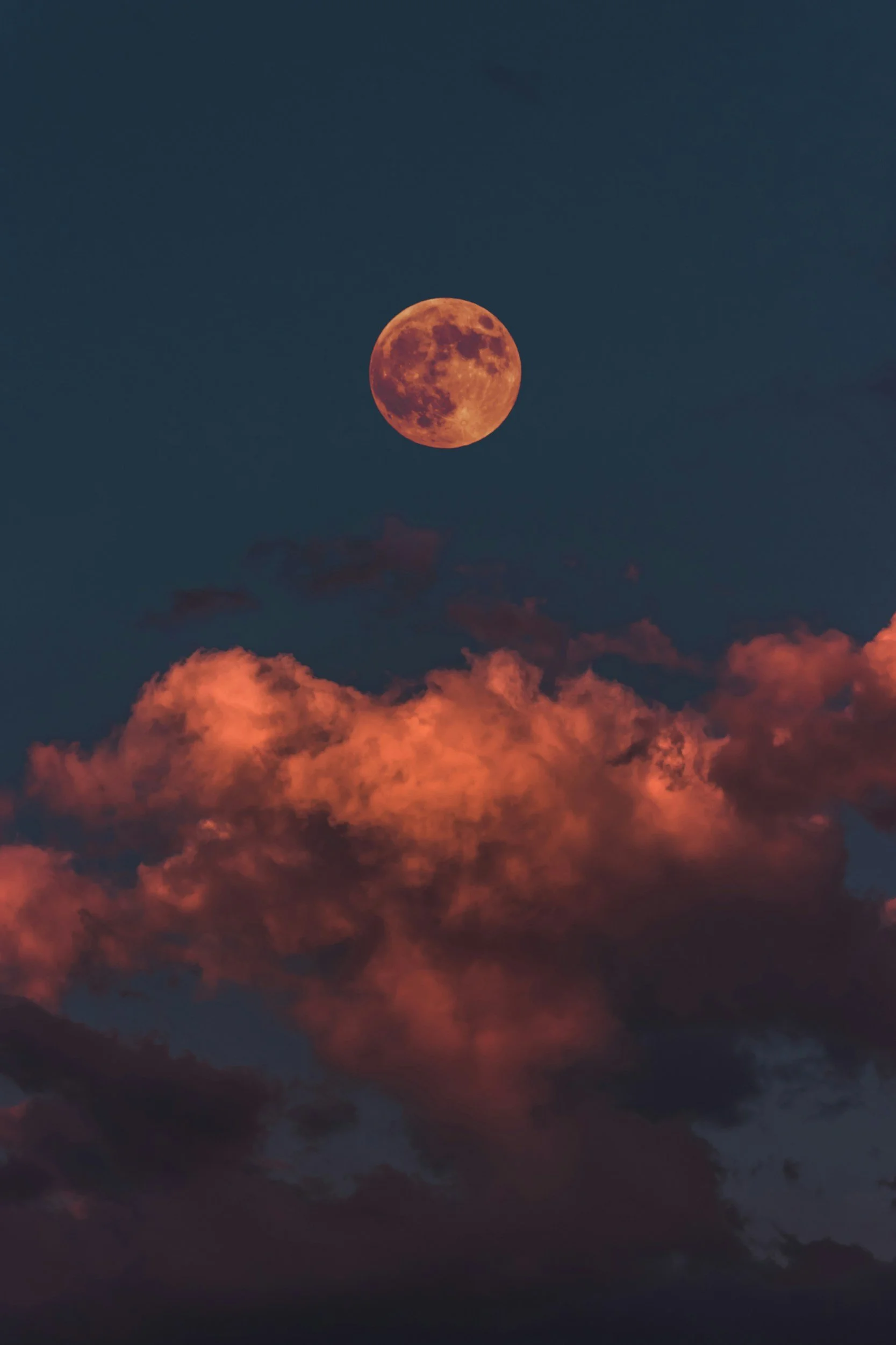 Der Mond leuchtet nachts in einem orange-roten Farbton am Himmel, umgeben von dunkelblauem Himmel und roten Wolken im Vordergrund.