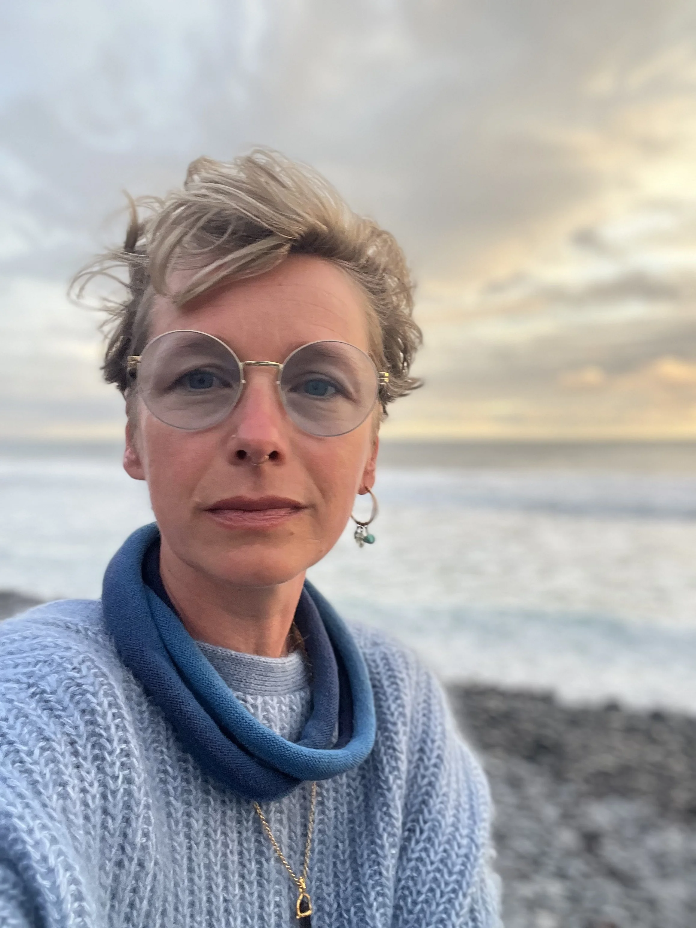 Nahaufnahme einer Frau mit kurzen, blonden, welligen Haaren und runden Sonnenbrillen vor einem Meer- oder Seelandschaft bei Sonnenuntergang, trägt einen grauen Pullover und eine blaue Jacke, trägt eine Halskette und einen Ohrring.