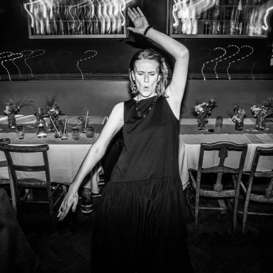 A woman dancing with her eyes closed and one arm raised in a black dress in a decorated dining area with chairs and vases filled with flowers.