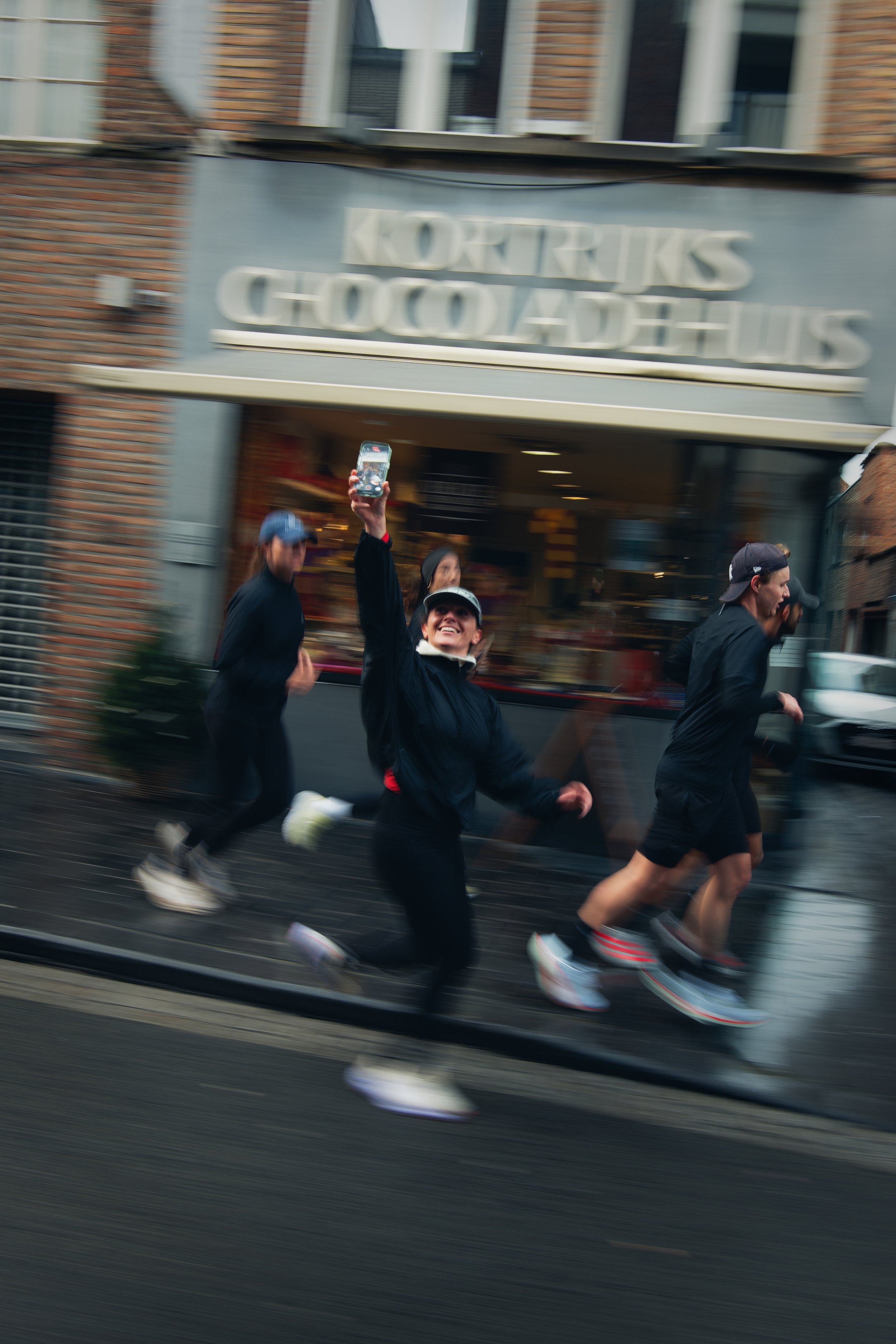 Mensen rennen op een straat, één persoon houdt een telefoon omhoog en lacht, in de achtergrond een winkel met een lichtbord.