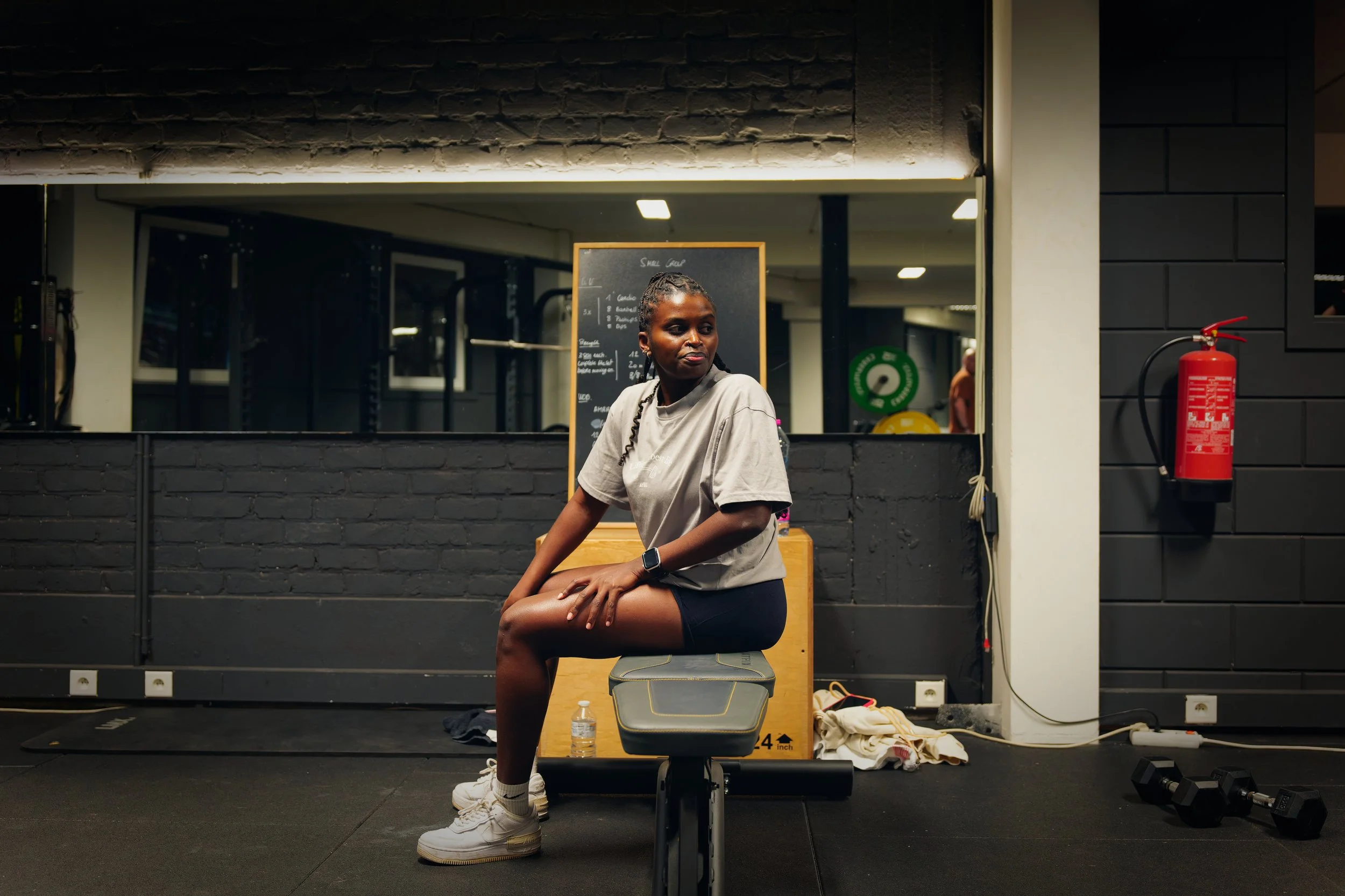 Een vrouw in een sportschool zit op een bankje voor een spiegel, met fitnessapparatuur en een waterfles op de grond naast haar.