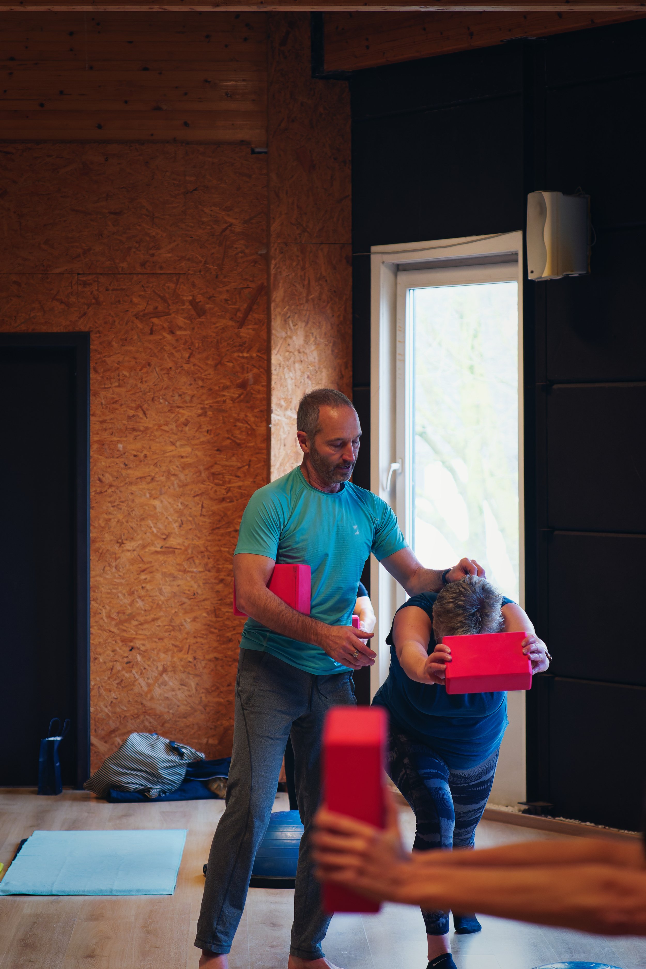 Een man helpt een vrouw met een oefening in een huiselijke omgeving, beiden maken gebruik van rode yoga blocks.