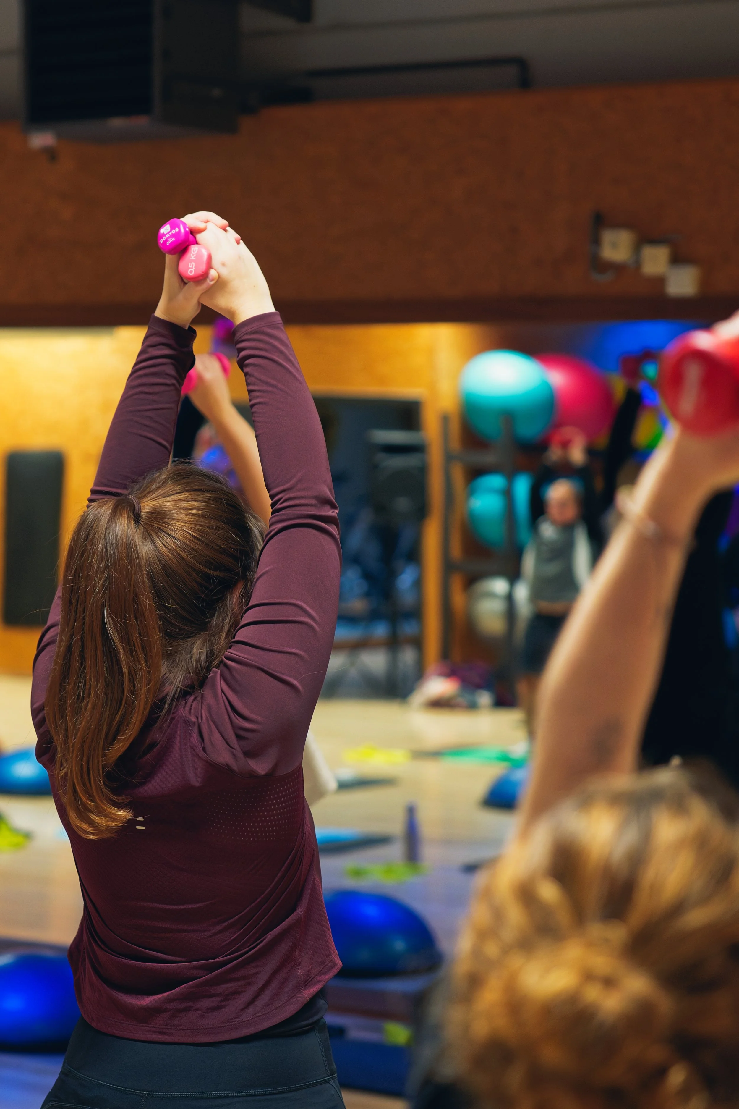 Glimlachende vrouwen in een fitnessstudio die deelnemen aan een groepsles, ondanks dat ze niet zichtbaar zijn in de foto, is de focus op een vrouw met bruin haar die een exercise met gewichten doet.