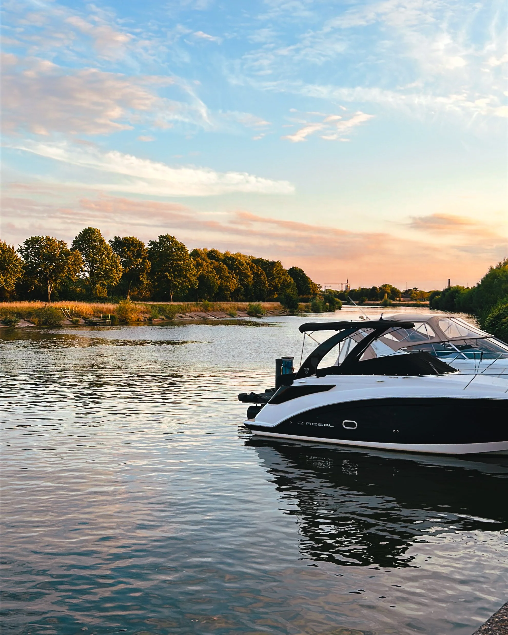 Een rivier met een zwart-wit speedboot en een groene bosachtige oever onder een blauwe, bewolkte hemel tijdens zonsondergang.