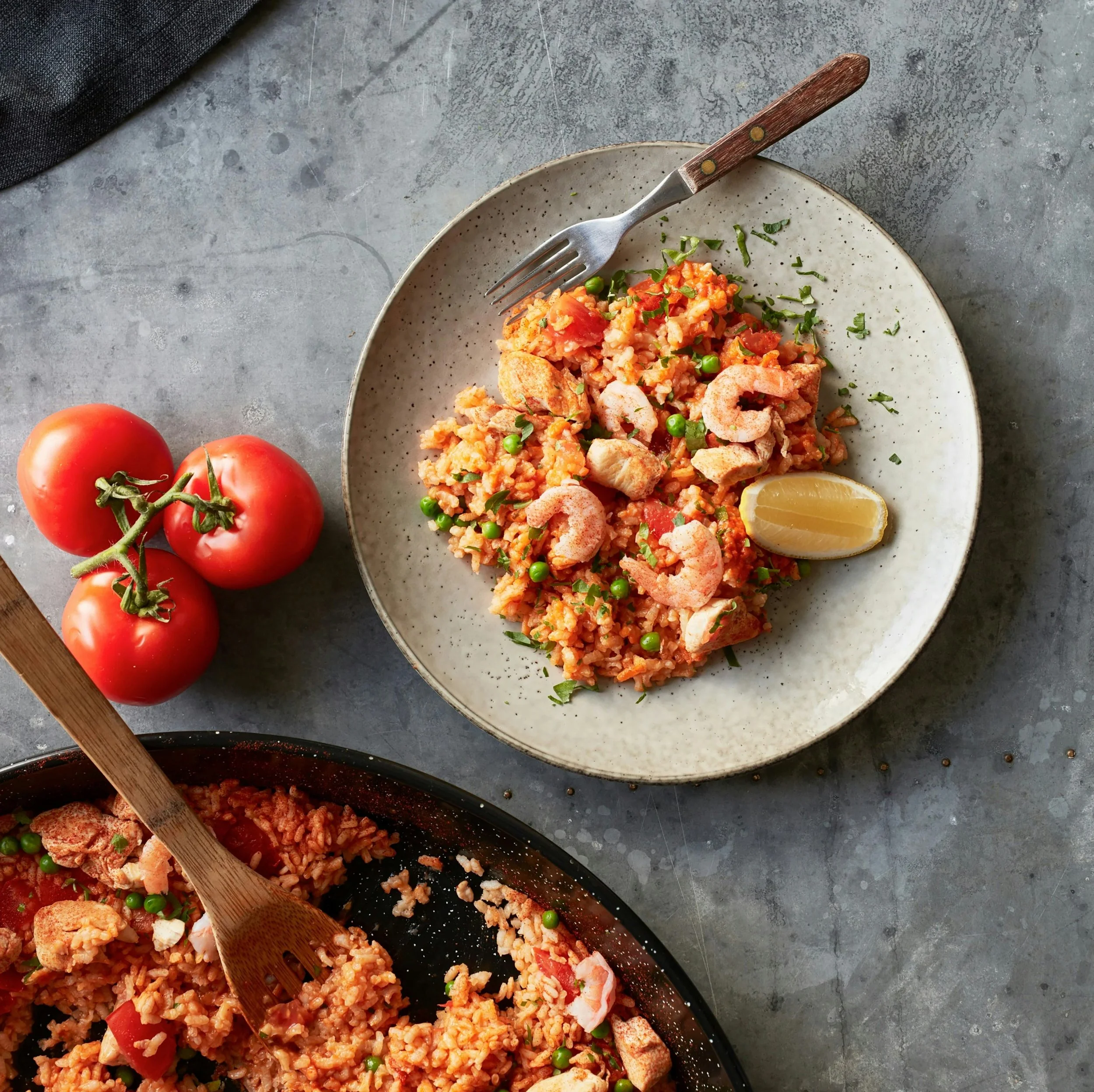 Gerecht met rijst, garnalen, groenten en een citroenpartje op een bord, naast tomaten en een schep met rijst in een pan.