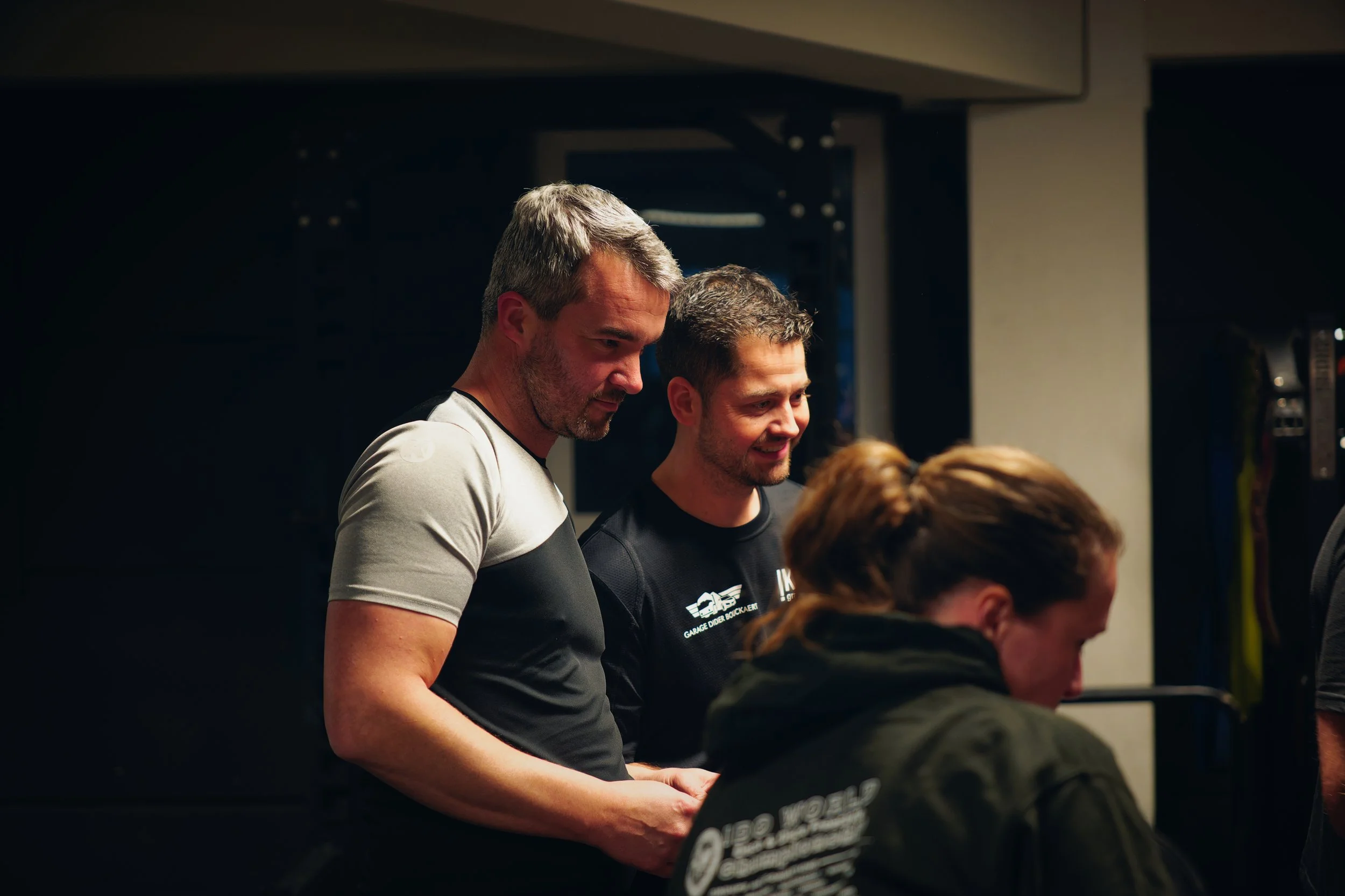 Drie mensen samen in een donkere kamer, ze kijken naar iets dat niet zichtbaar is op de foto. Twee mannen staan dicht bij elkaar, een vrouw zit met haar rug naar de camera.