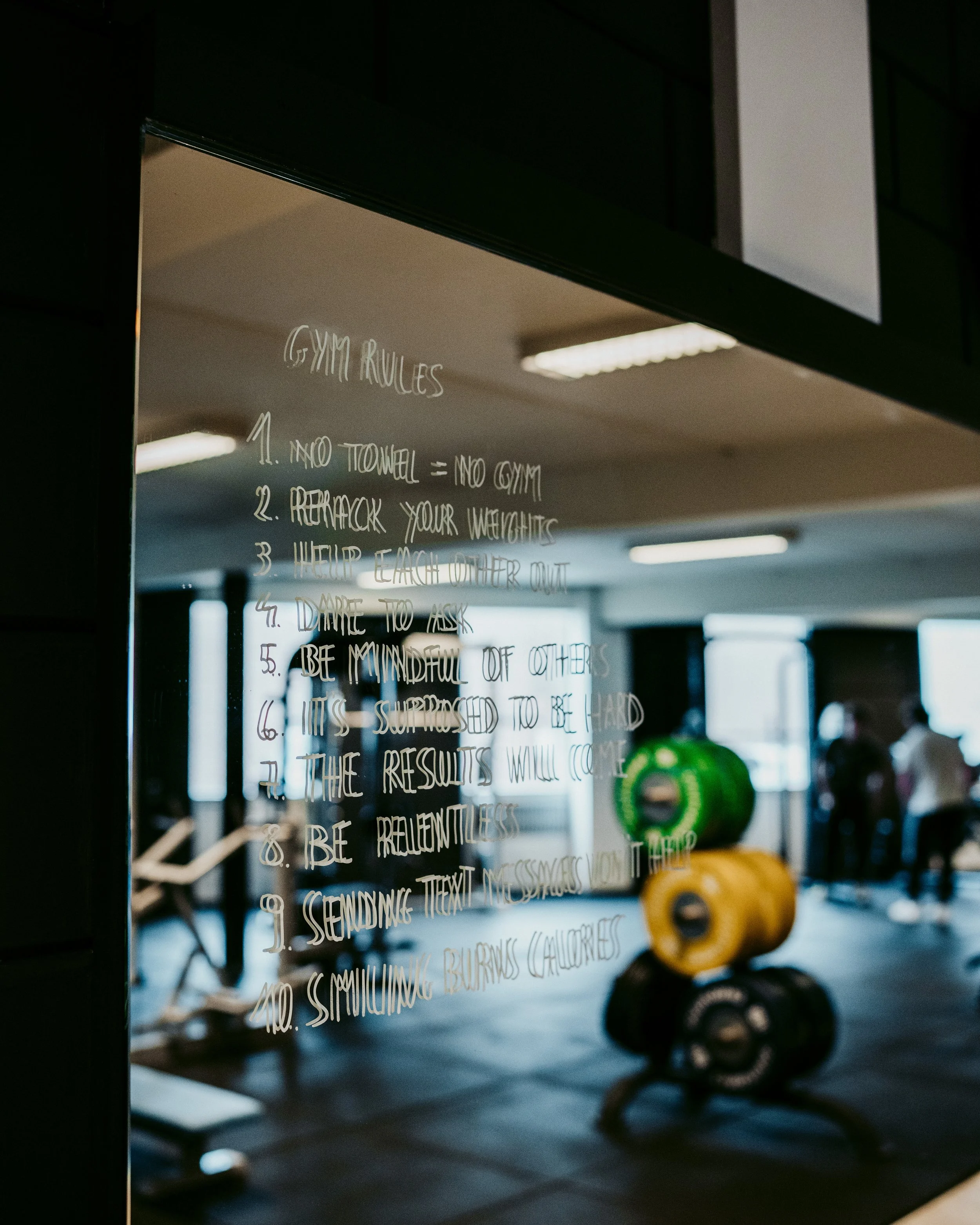 Een spiegel met fitnessregels geschreven erop, met in de achtergrond gewichten en mensen aan het sporten in een sportschool.