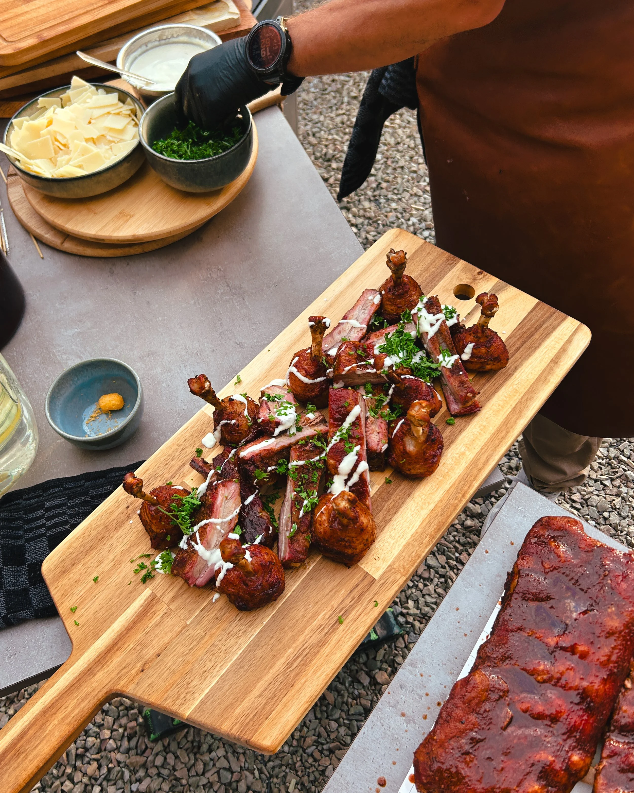 Gegrilde vleesgerechten zoals spareribs en kippoten op een houten serveerbord, versiering met peterselie, op een buitentafel met verschillende sauzen en kruiden.