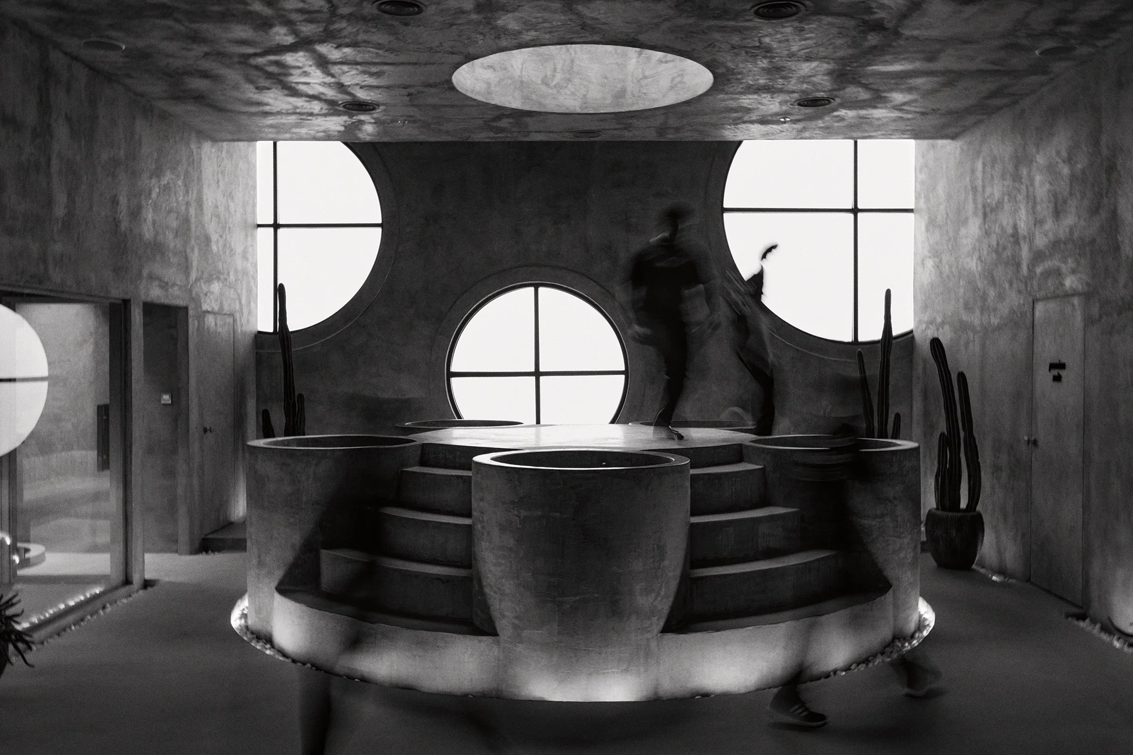 Binnenruimte met grote ronde ramen, trap en planten, silhouetten van twee mensen die dansen.