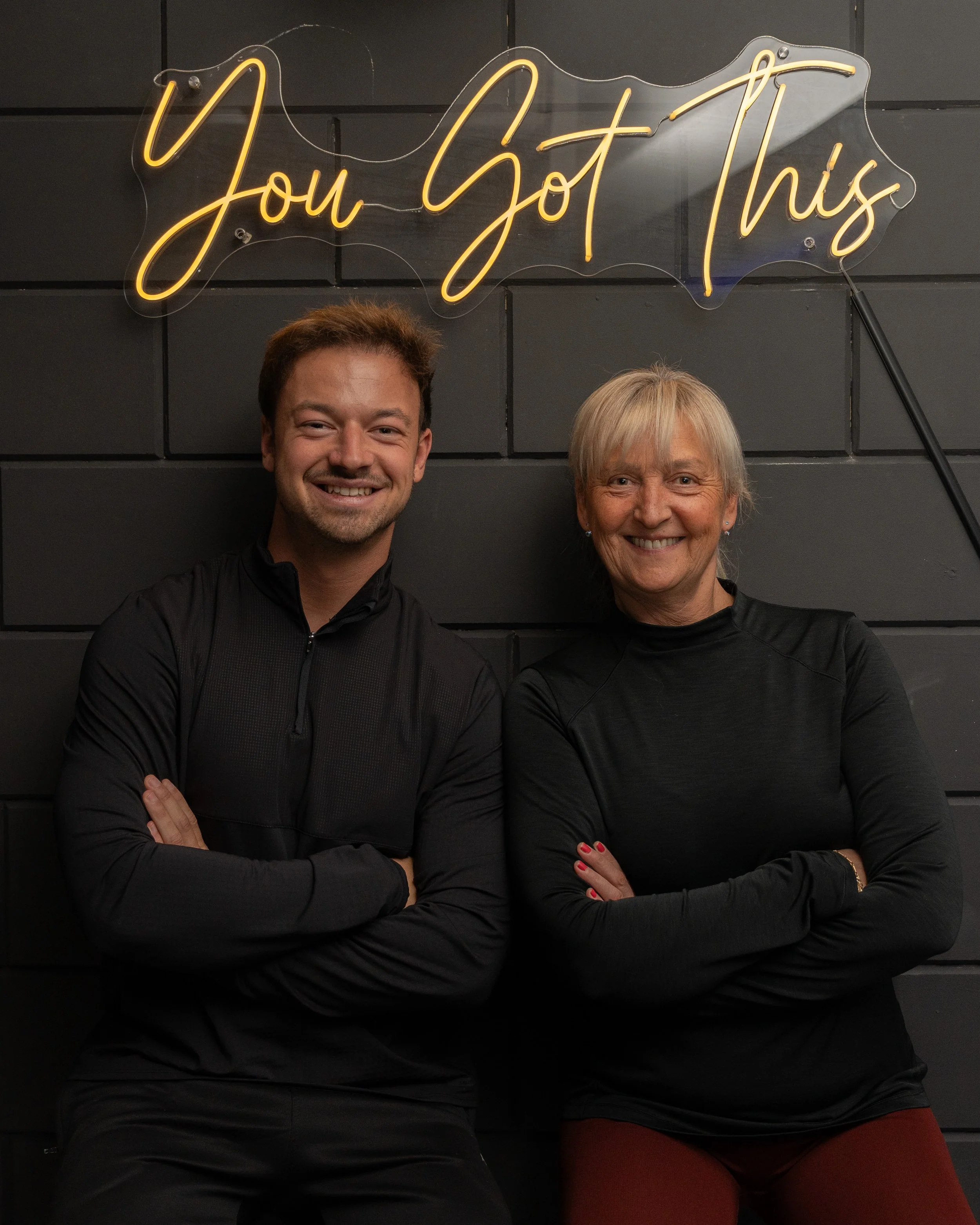 Een man en een vrouw staan lachend voor een zwarte muur met een verlichte neonboodschap die zegt 'You Got This'.