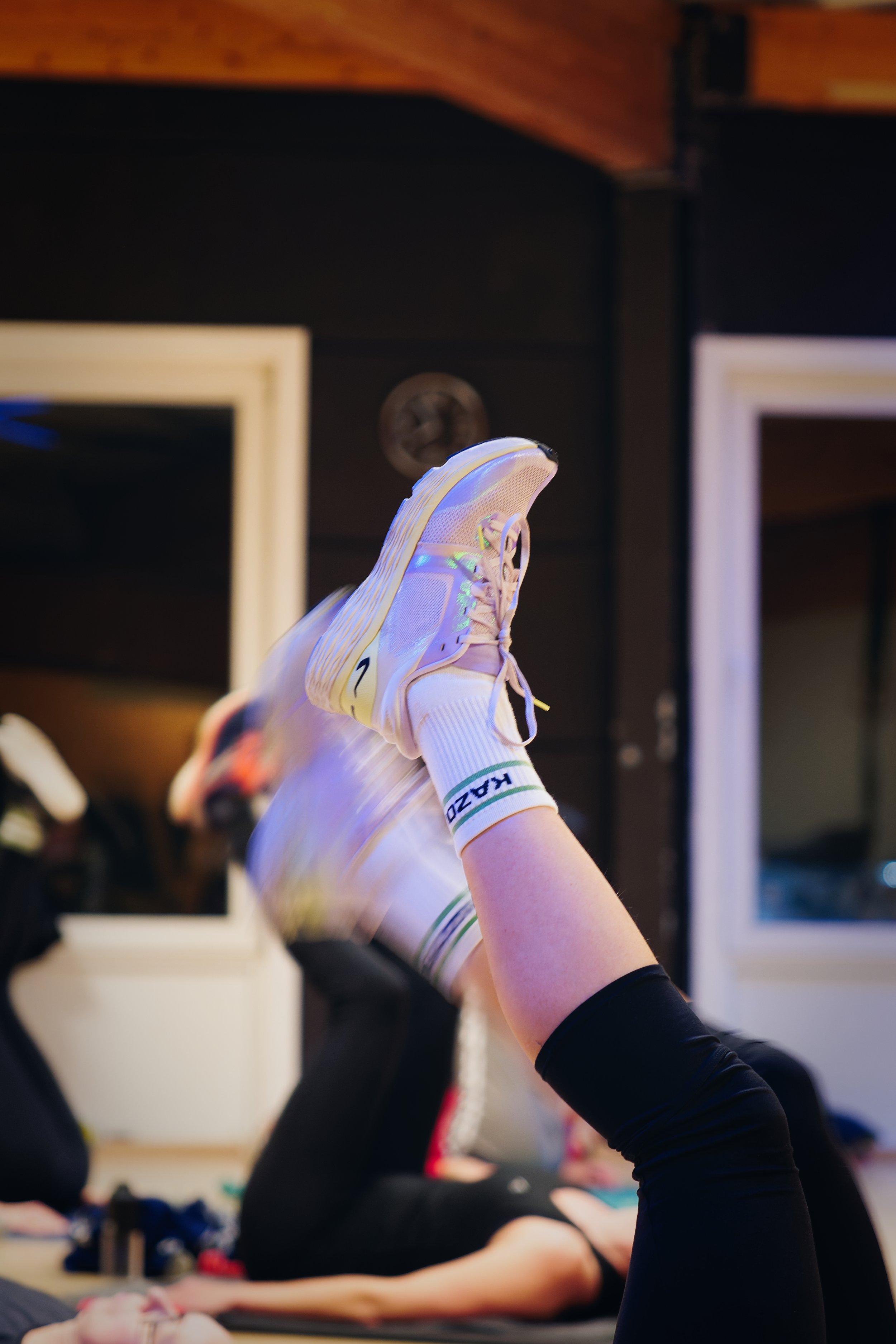 Persoon die in een yogapose ligt met de benen omhoog, draagt witte sportschoenen en witte sokken met groene en blauwe details, in een binnensituatie.