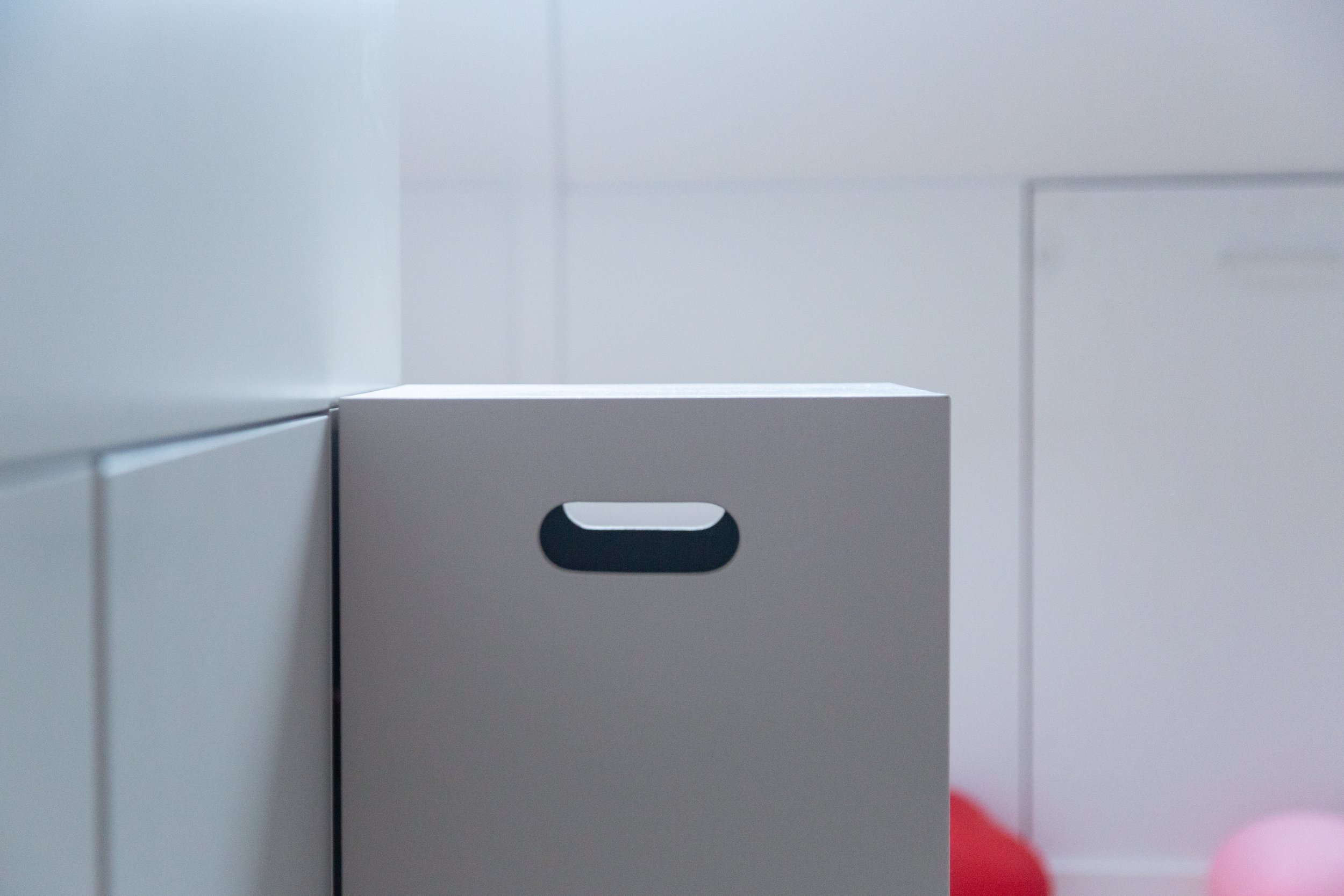 Een witte lade kast met een open vak erin, in een minimalistische kamer met wit wand en vloer, met een paar ballonnen op de vloer op de achtergrond.