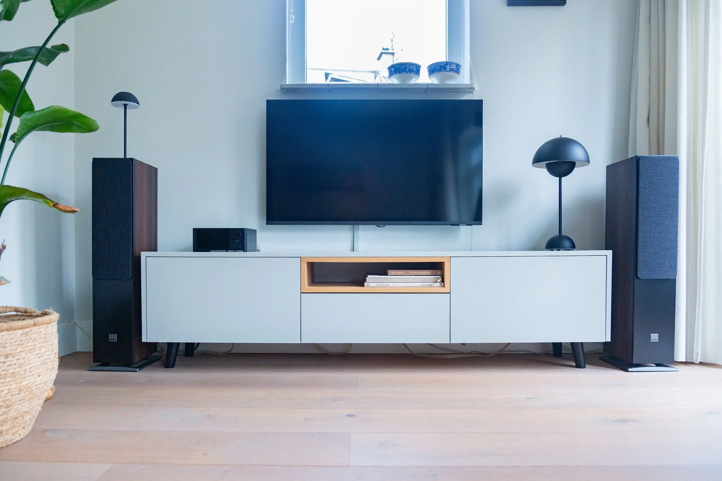 Mooie woonkamer met witte tv-meubel, twee zwarte grote luidsprekers aan weerszijden, een grote platte tv op de muur, een kleine zwarte audio-apparaat, twee moderne zwart lampen, een venster met gordijnen, en een groene plant in een mand.