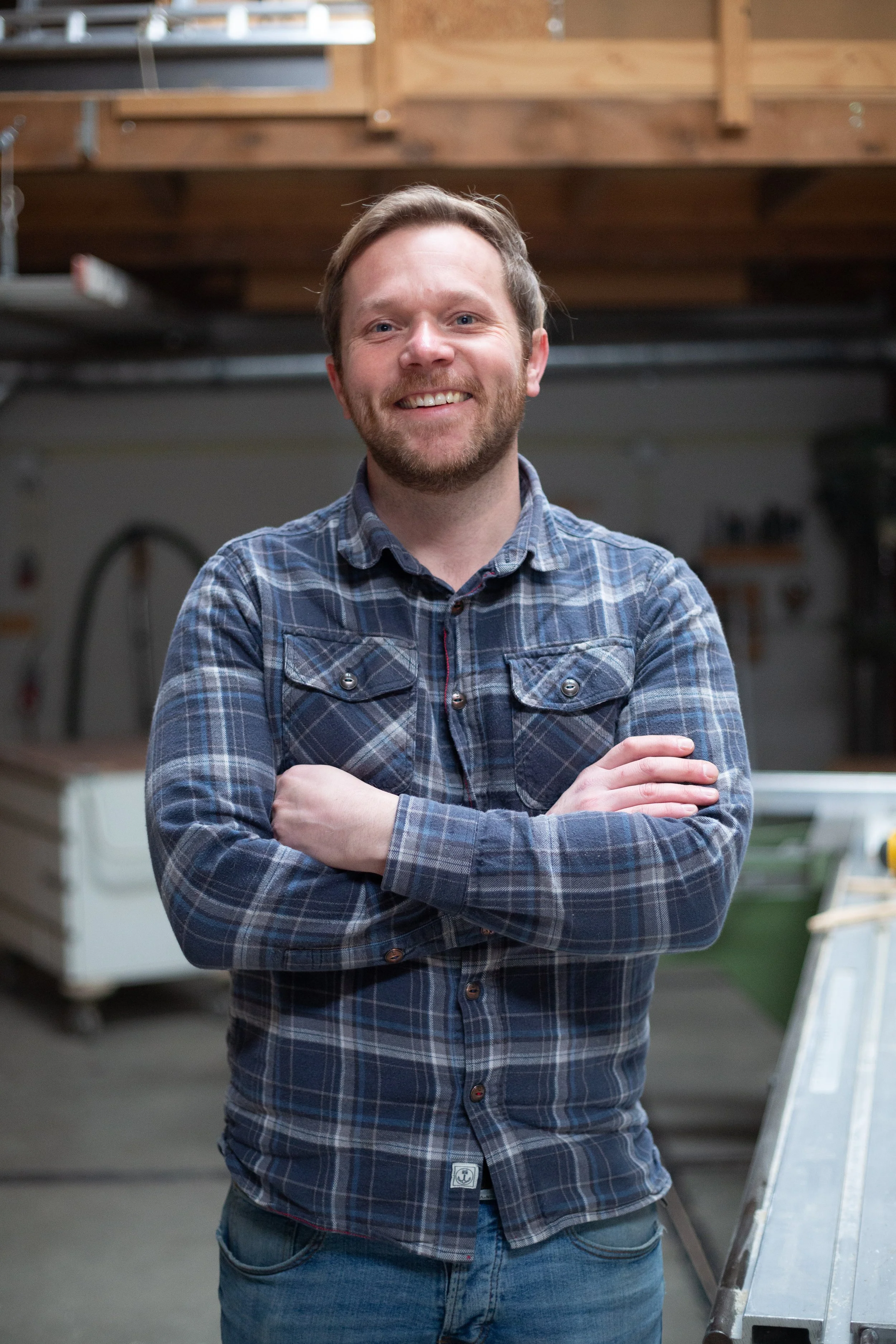 Een lachende man met een baard en kort haar, gekleed in een blauw geruit overhemd, staat met gekruiste armen in een werkplaats of garage.