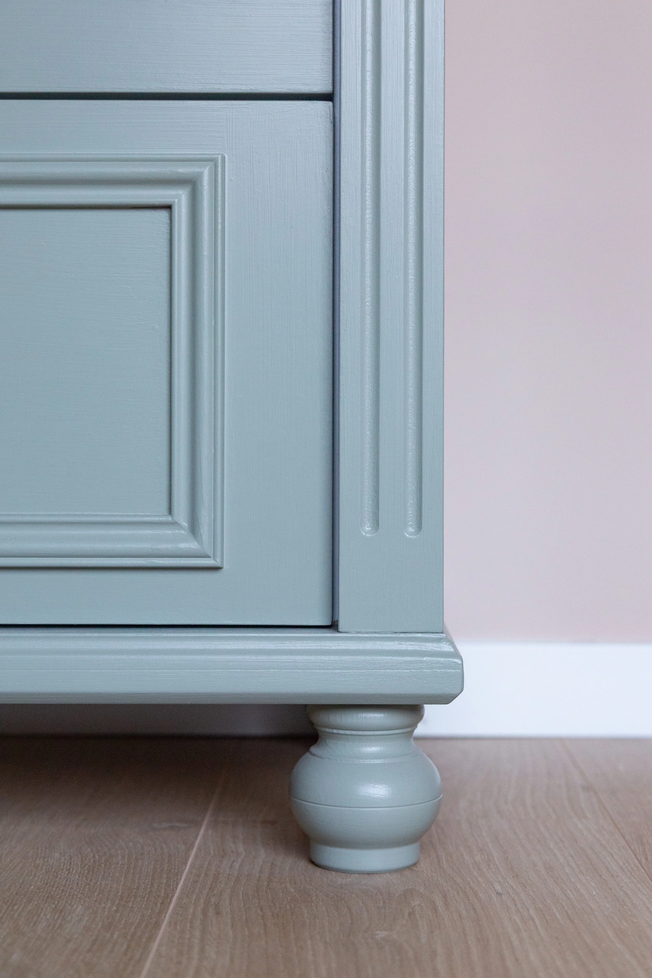 Close-up van een lichtblauwe houten meubelkast met een gedraaide poot, op een houten vloer en een zachte roze muur op de achtergrond.