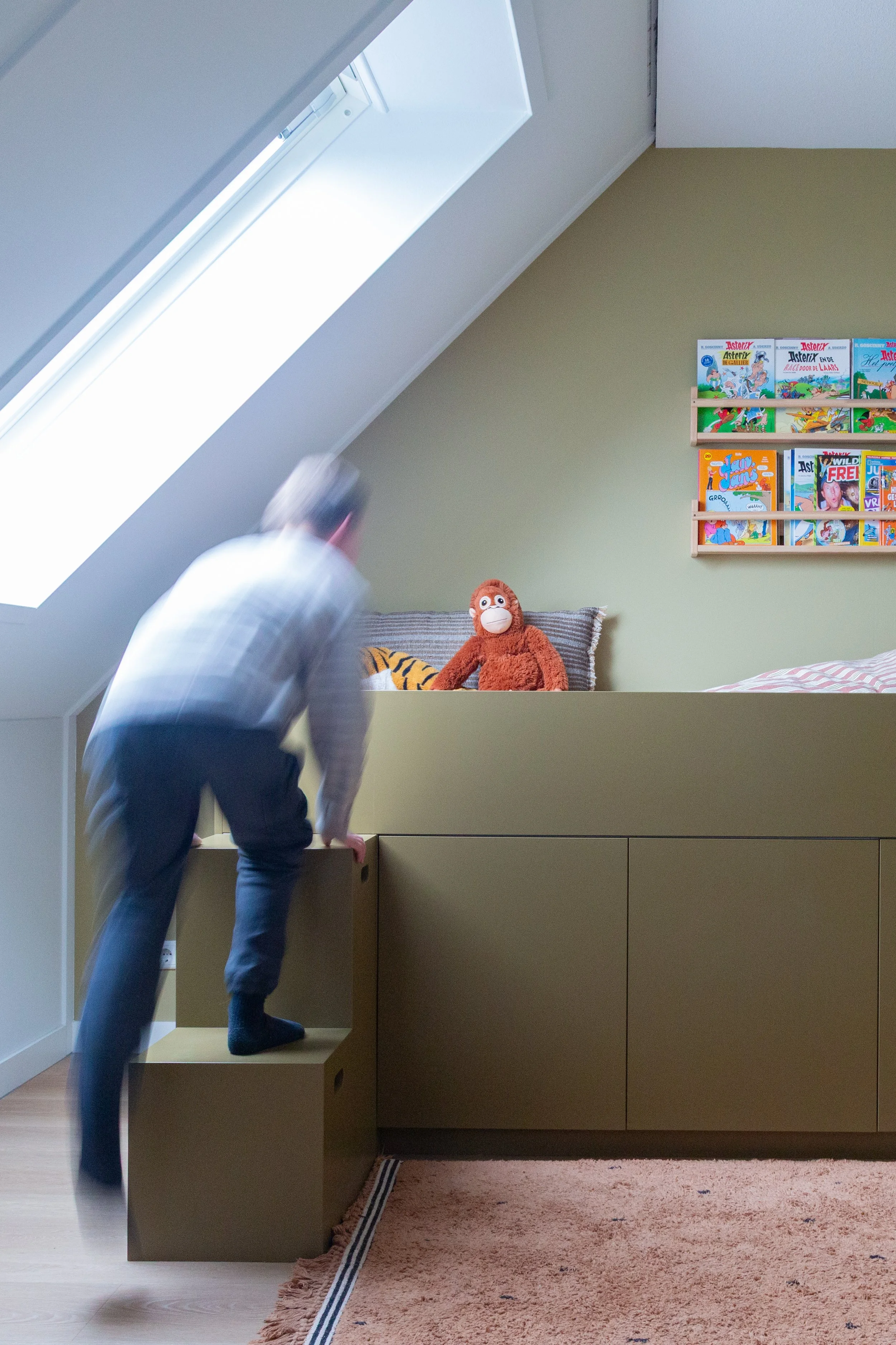 Een jongen klimt op een houten trapje naar een hoogslaper in een kinderkamer met een gebreide deken en knuffels, waaronder een aap en een tijger, op het bed. Op de muur hangt een boekenrek met kinderboeken.