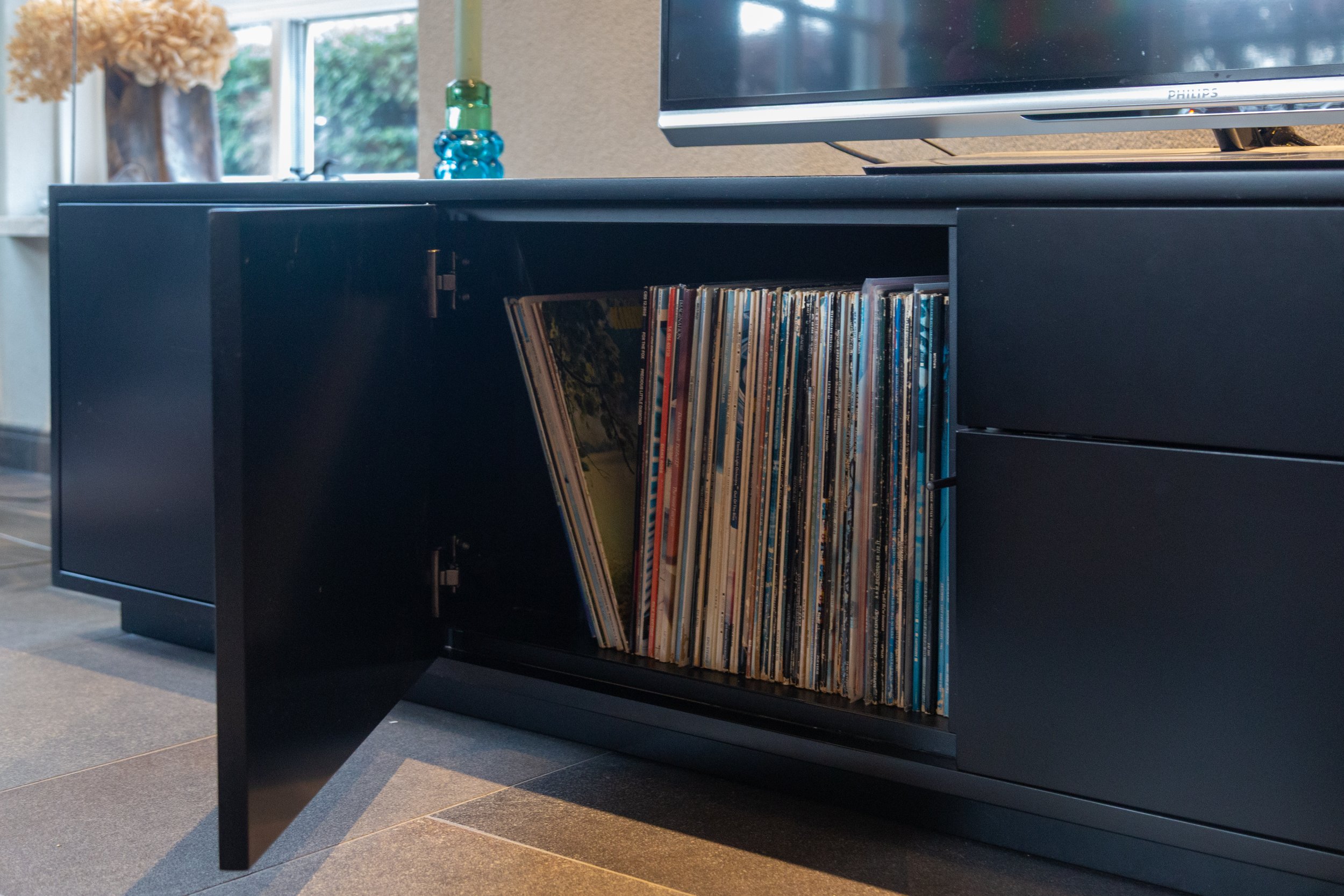 Een zwarte lage kast met een open deur, gevuld met vinylplaten in een woonkamer met een tv erboven en decoratieve voorwerpen op de tafel naast een raam.