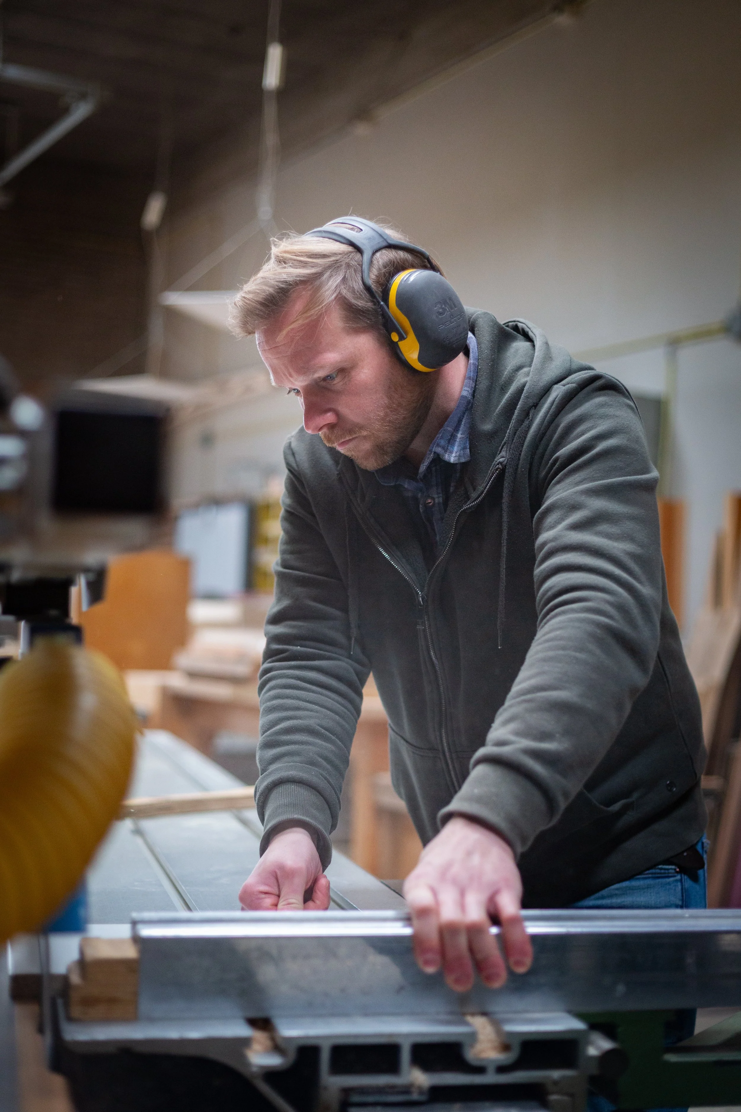 Man met oorbeschermers die werkt in een houtwerkplaats en een stuk hout bewerkt met een zaag.