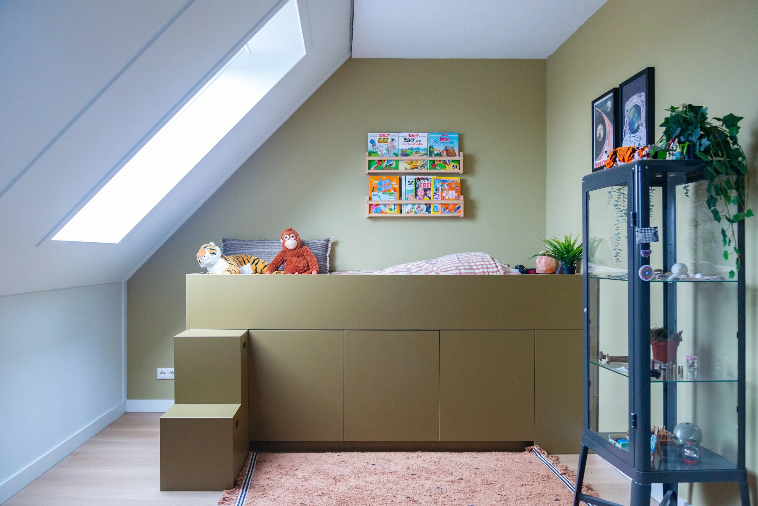 Een kinderkamer met een bed onder een schuin dak en een grote raam, decoratie met knuffels en speelgoed, een glazen kast met speelgoed en planten, muurdecoraties, en een rustgevende, georganiseerde uitstraling.