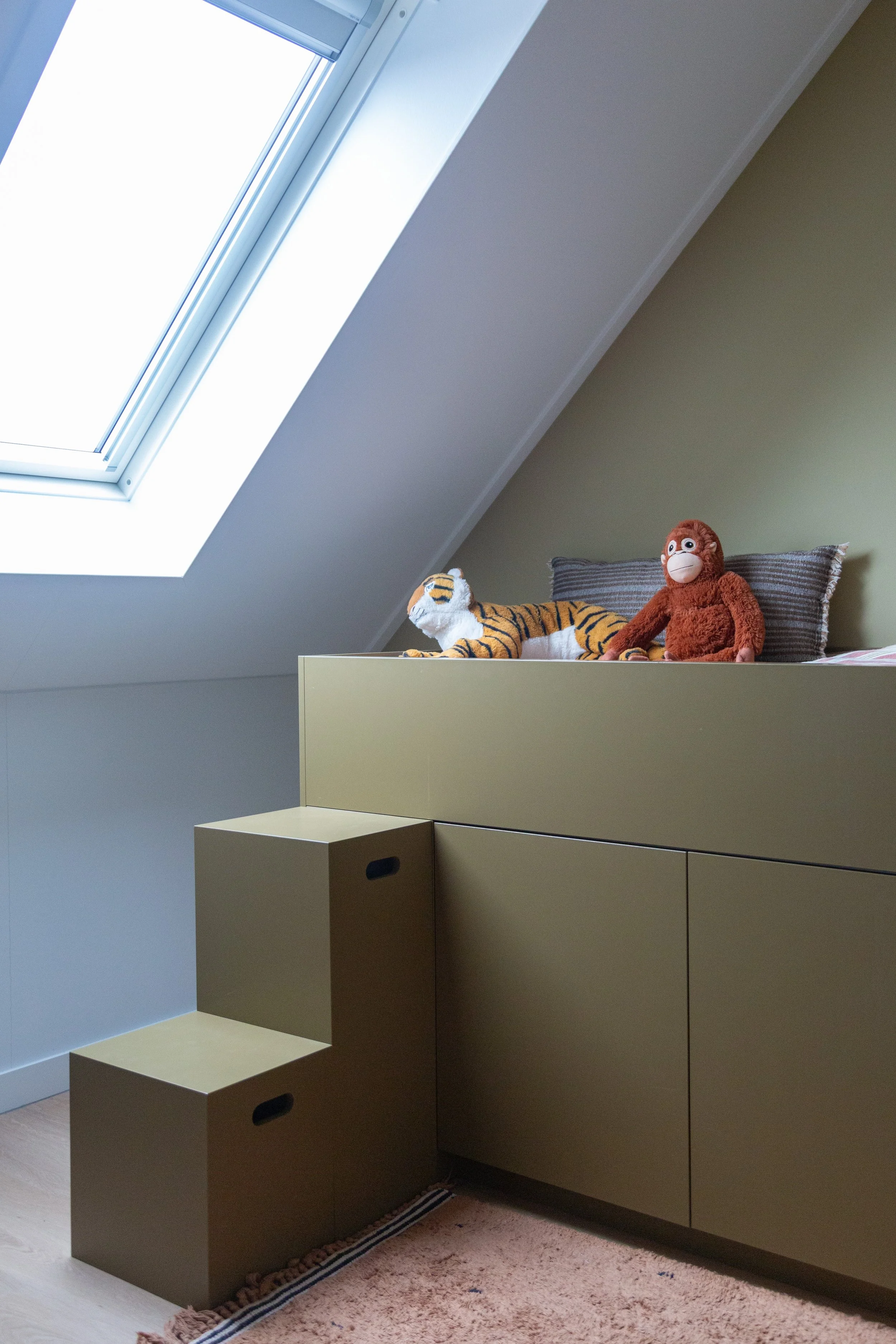 Een slaapkamer met een schuine dakramen, een zwarte decoratieve muur, en op een beige kast staan twee knuffeldieren: een tijger en een orang-oetan, met een kussen op de kast.