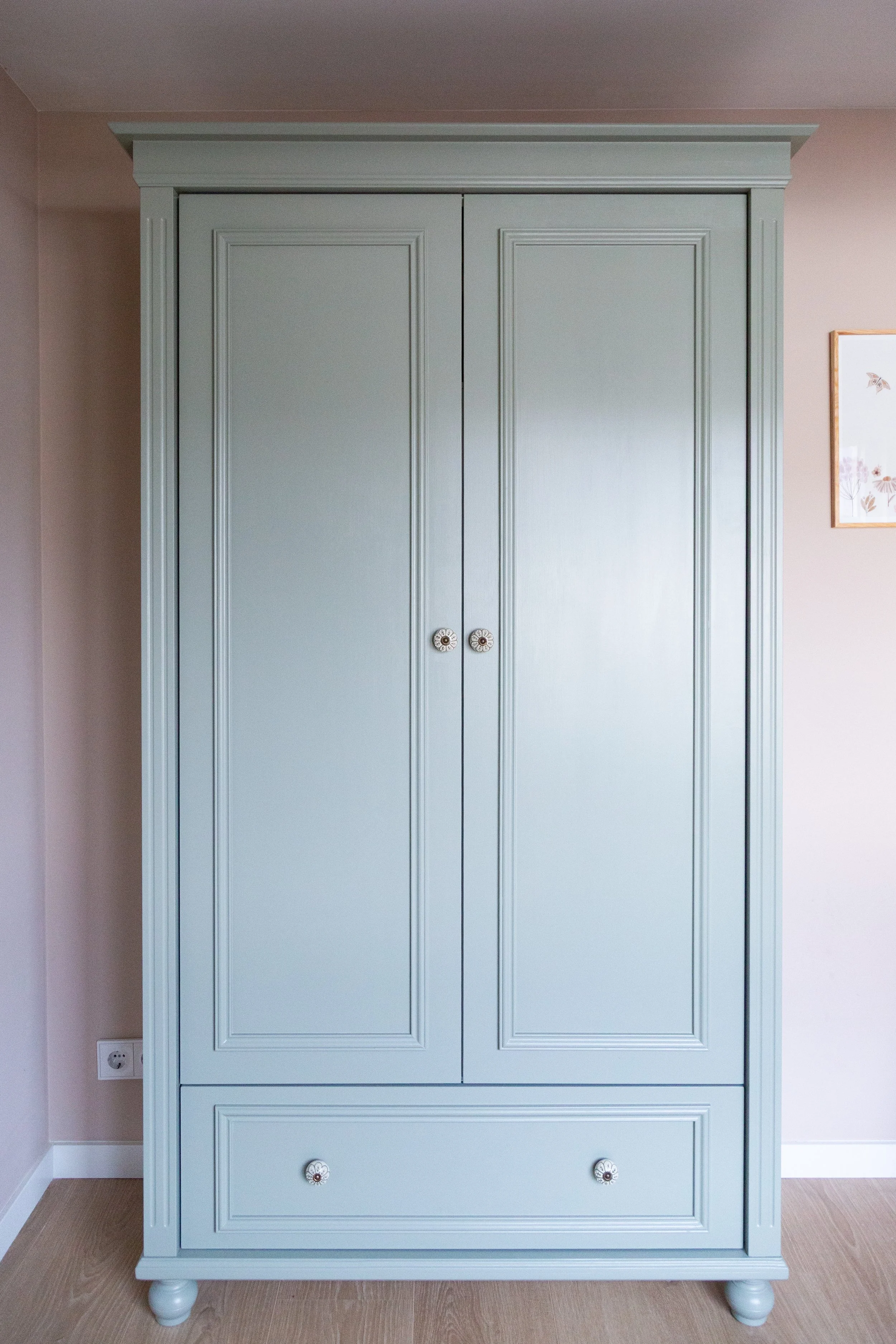 Lichtblauwe houten kast met twee deuren en een lage lade onderaan, geplaatst tegen een lichte muur in een interieur. Op de muur hangt een ingelijste botanische print.