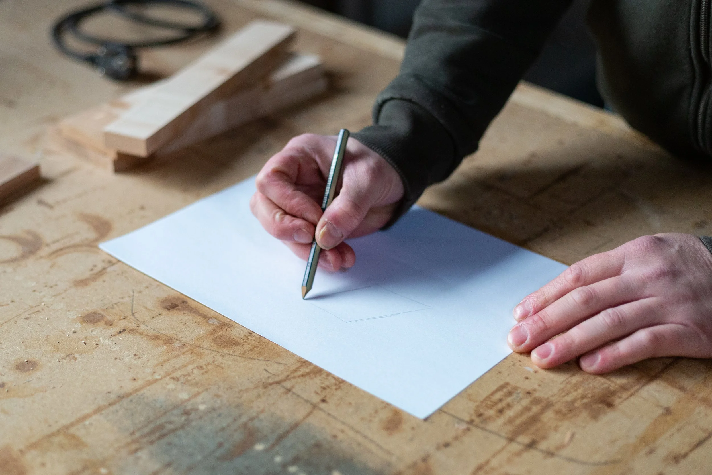 Persoon tekent op een stuk wit papier met potlood op een houten werkbank, met gereedschap en hout op de achtergrond.