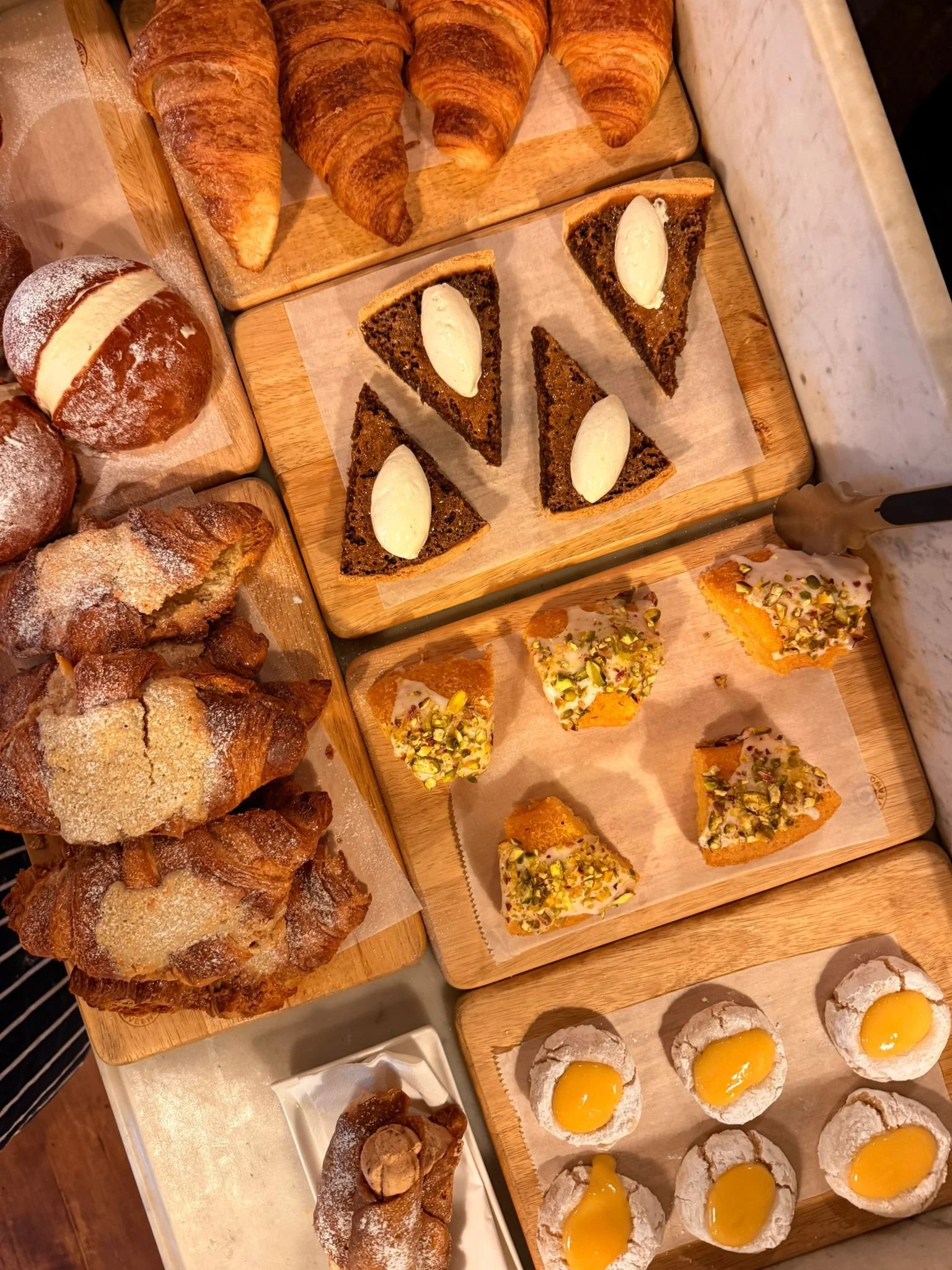 An assortment of baked goods on wooden trays, including croissants, round bread rolls, cruffins, triangular slices of nut-topped pastry, triangular slices of chocolate cake with cream, and small desserts with yellow filling, possibly lemon tarts or cookies with egg yolk.