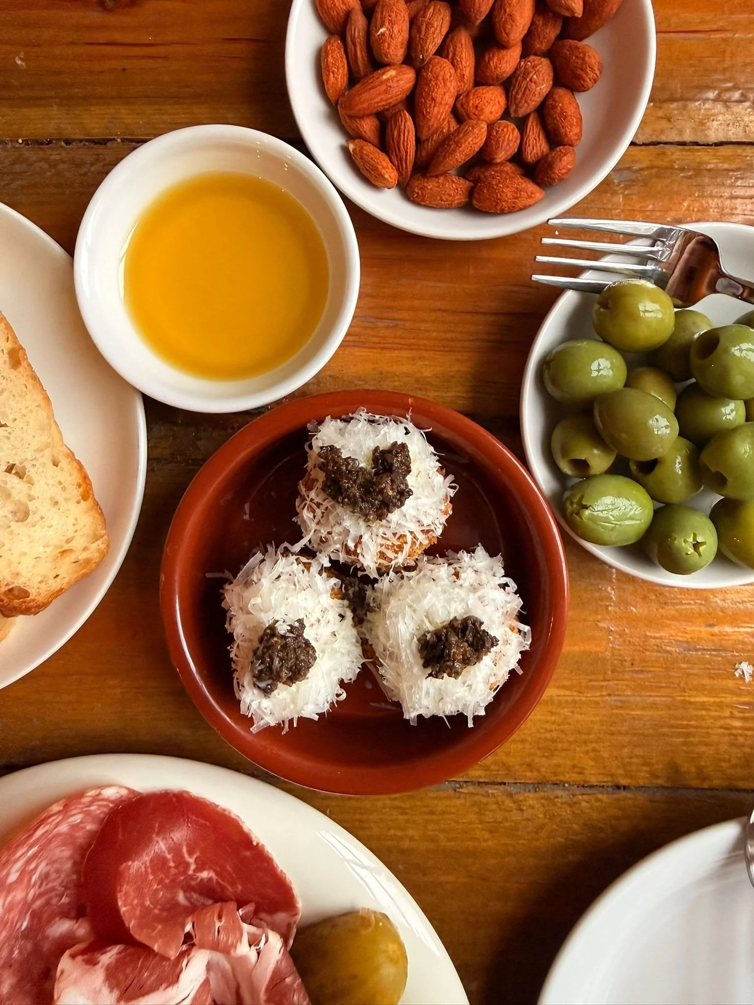 An assortment of Mediterranean dishes on a wooden table, including a bowl of almonds, green olives, a small bowl of olive oil, slices of bread, cured ham, and three small servings of a cheese and eggplant dish topped with shredded cheese and black paste.