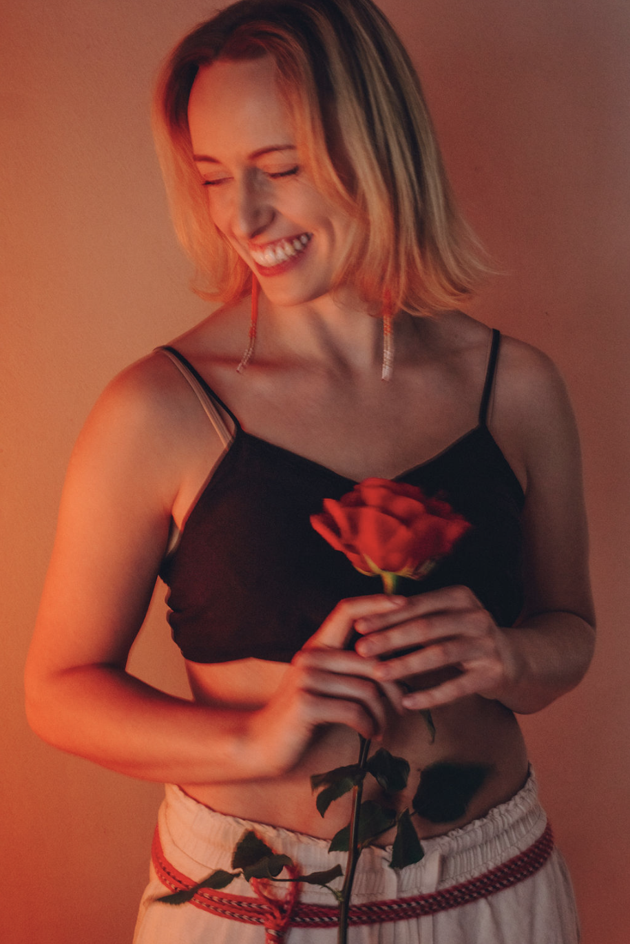 Lühikeste blondide juustega naine, kes naeratab ja hoiab käes punast roosi, kannab musta varrukateta pluusi ja heledaid pükse, seistes sooja värvi taustal.