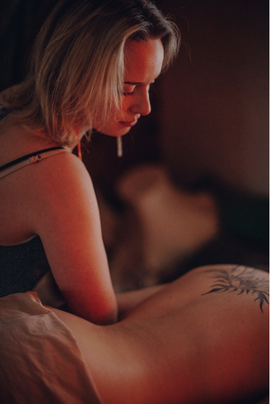 Lähivõttes sooja valgusega siseruumis on blondide juustega, musta pluusi ja kõrvarõngastega naine, kes kummardub ettepoole ja masseerib kliendi tätoveeritud selga.