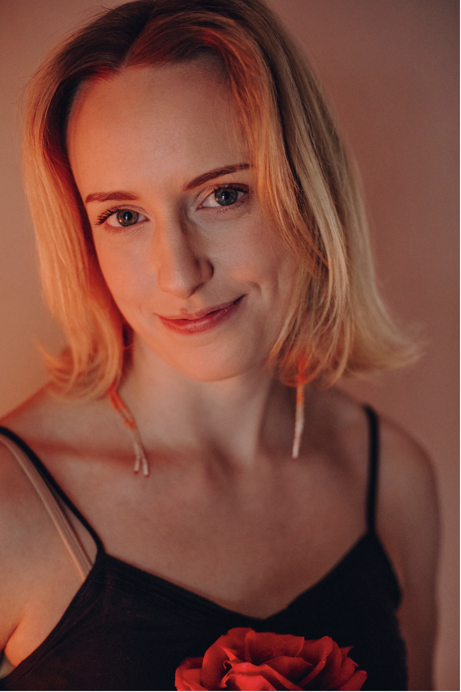 Õlgadeni laineliste blondide juustega naine, seljas must spagetipaeltega topp, õrnalt naeratav, käes punane roos.