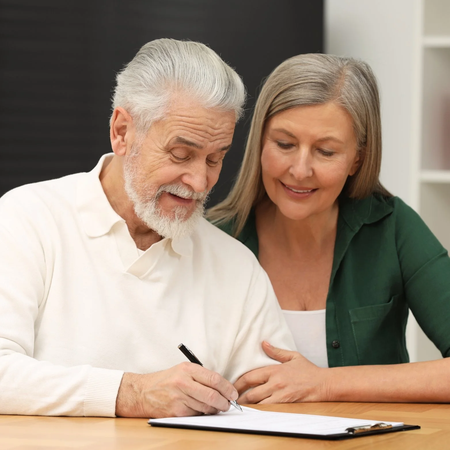 An elderly man with white hair and a beard signing a document while a woman stands beside him, smiling.