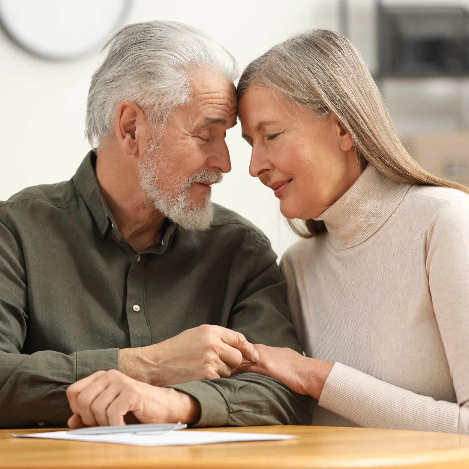 An elderly man and woman sitting close with foreheads touching and eyes closed, holding hands and smiling softly.