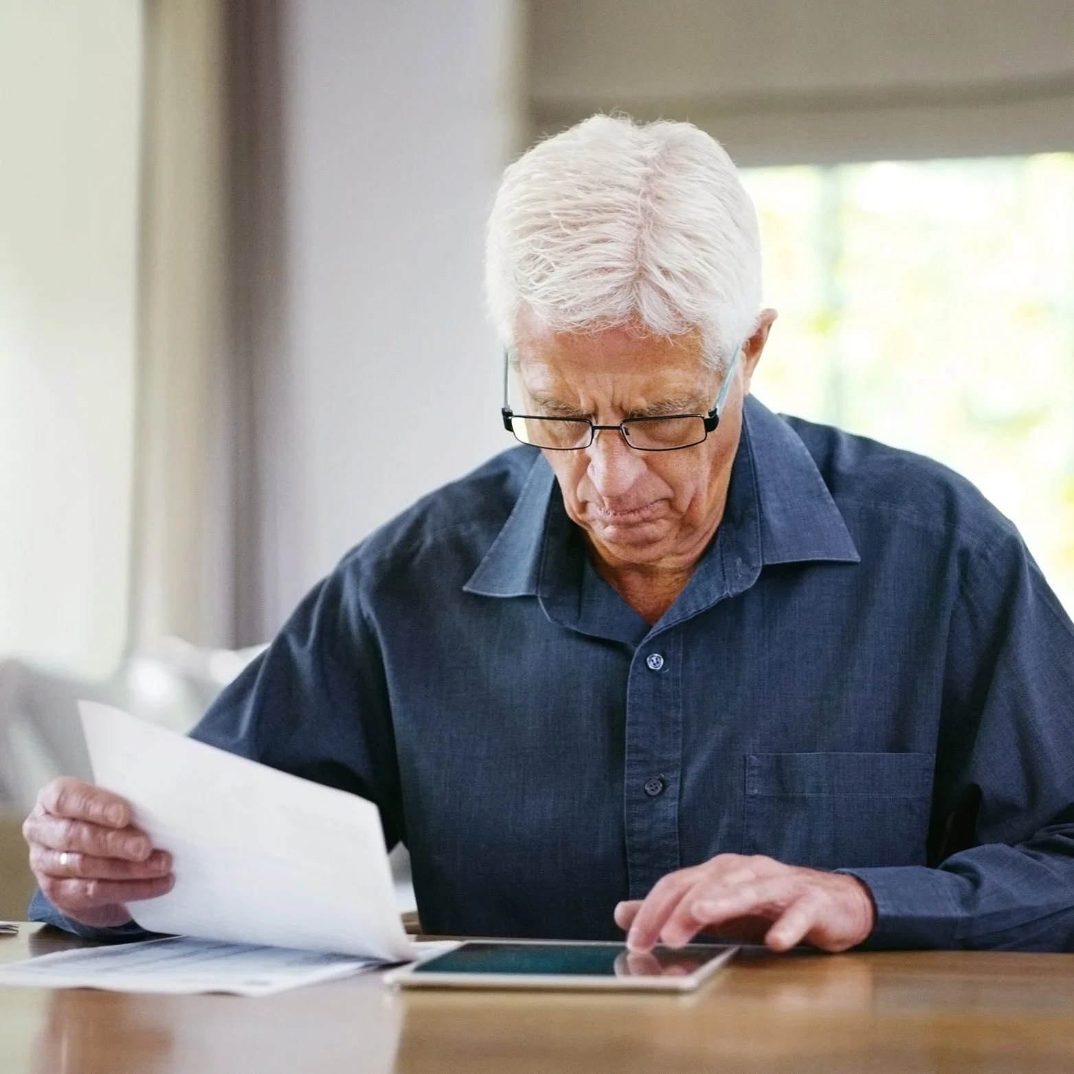 An elderly man with white hair and glasses wearing a dark blue button-up shirt sitting at a table, holding a piece of paper and looking at a tablet with a serious expression.