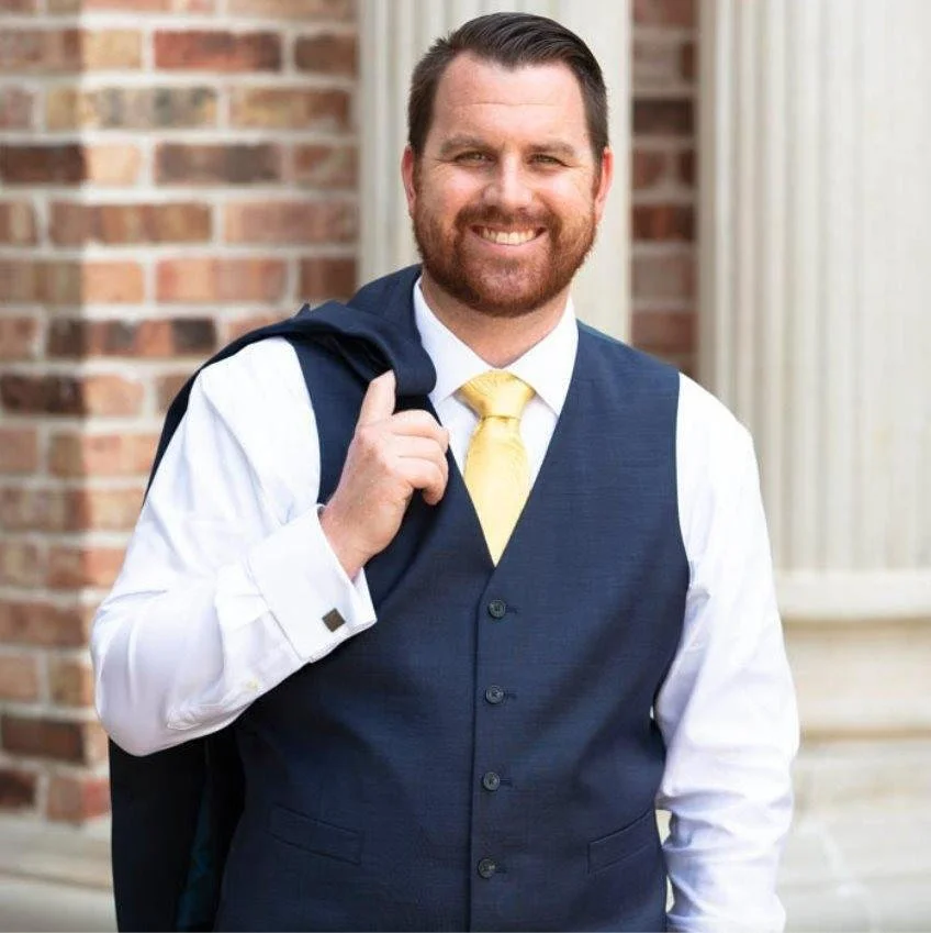 A man with a beard in a white shirt, navy vest, and yellow tie standing outside in front of a brick building, smiling, carrying a navy backpack on his shoulder.