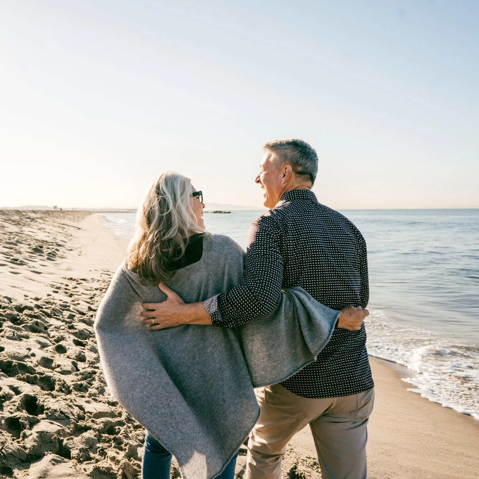 A senior couple walking on the beach at sunset, smiling and enjoying each other's company.