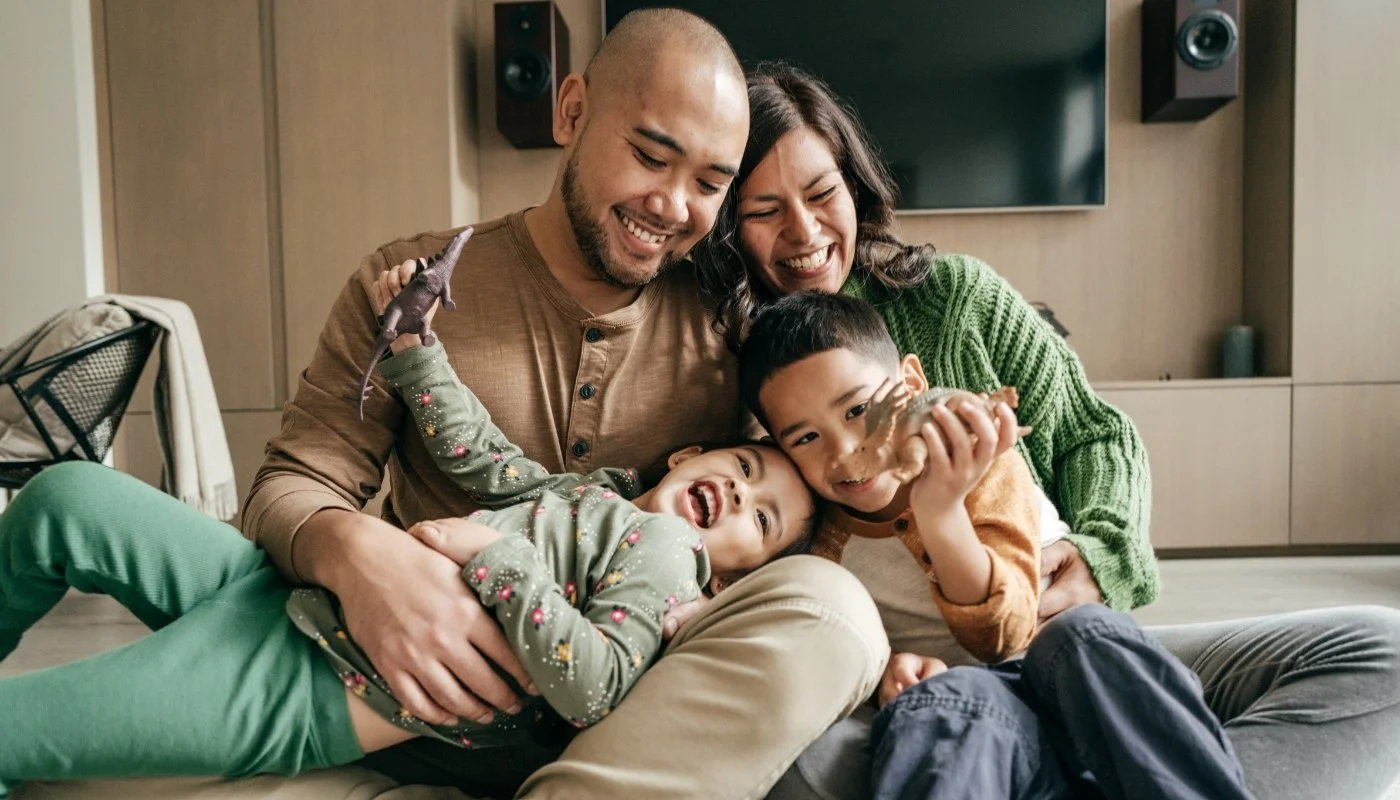 Smiling family sitting on the floor together, holding a toy dinosaur, in a cozy living room.