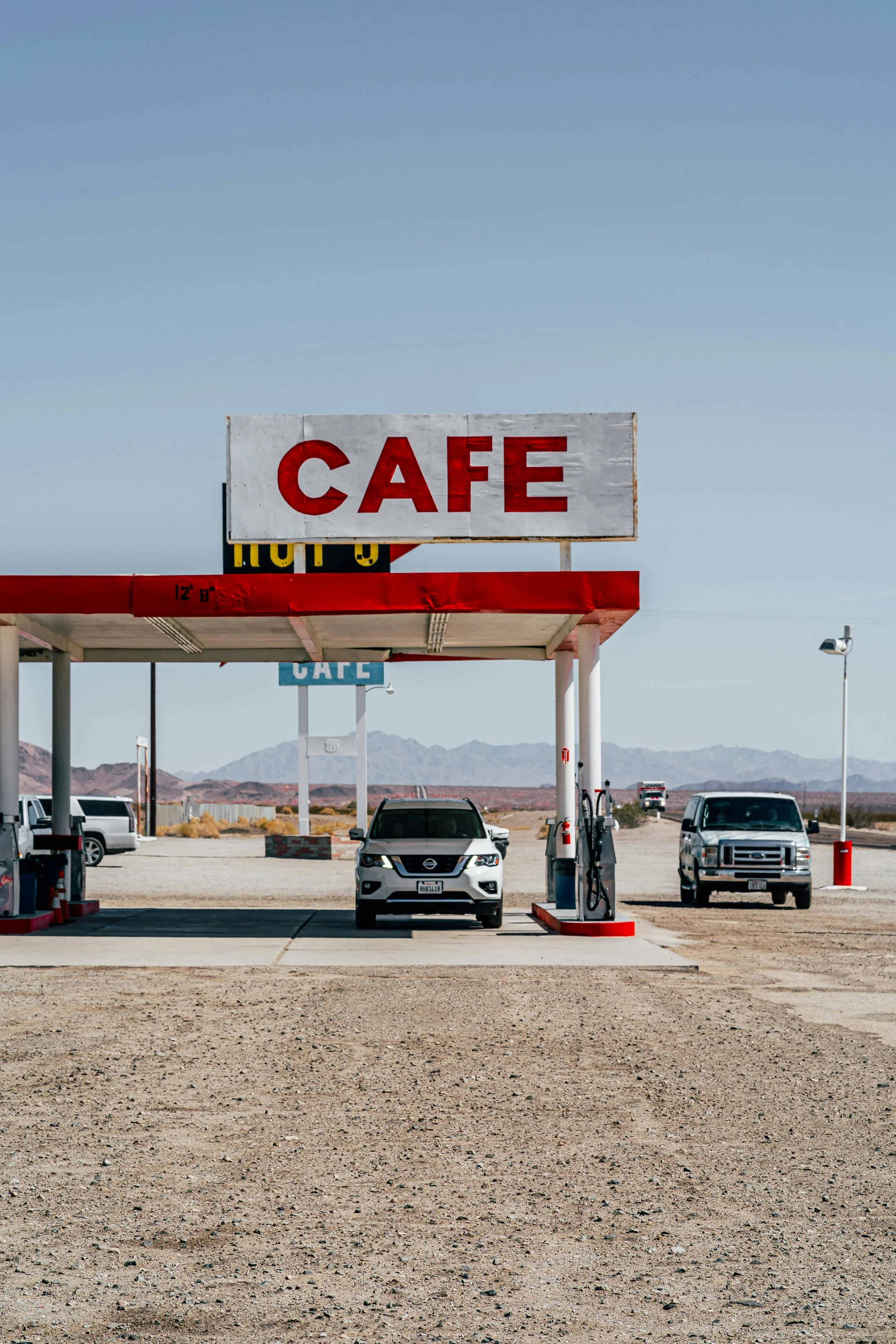 Station-service avec un véhicule à l'essence sous un panneau 'Café', dans un paysage désertique avec montagnes en arrière-plan.
